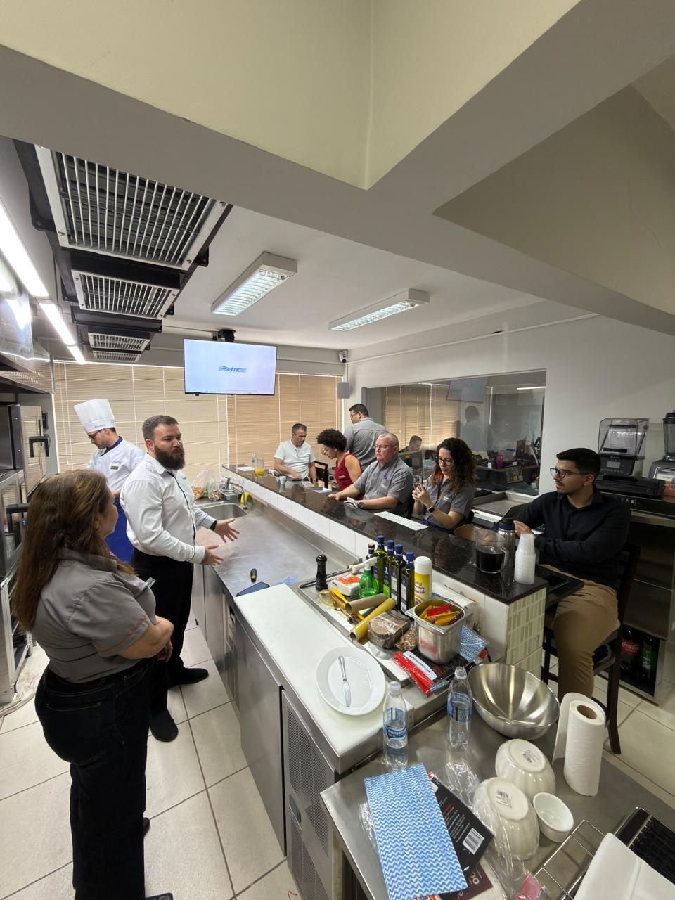 Pessoas na cozinha; um chef faz uma apresentação. Outros observam no balcão.