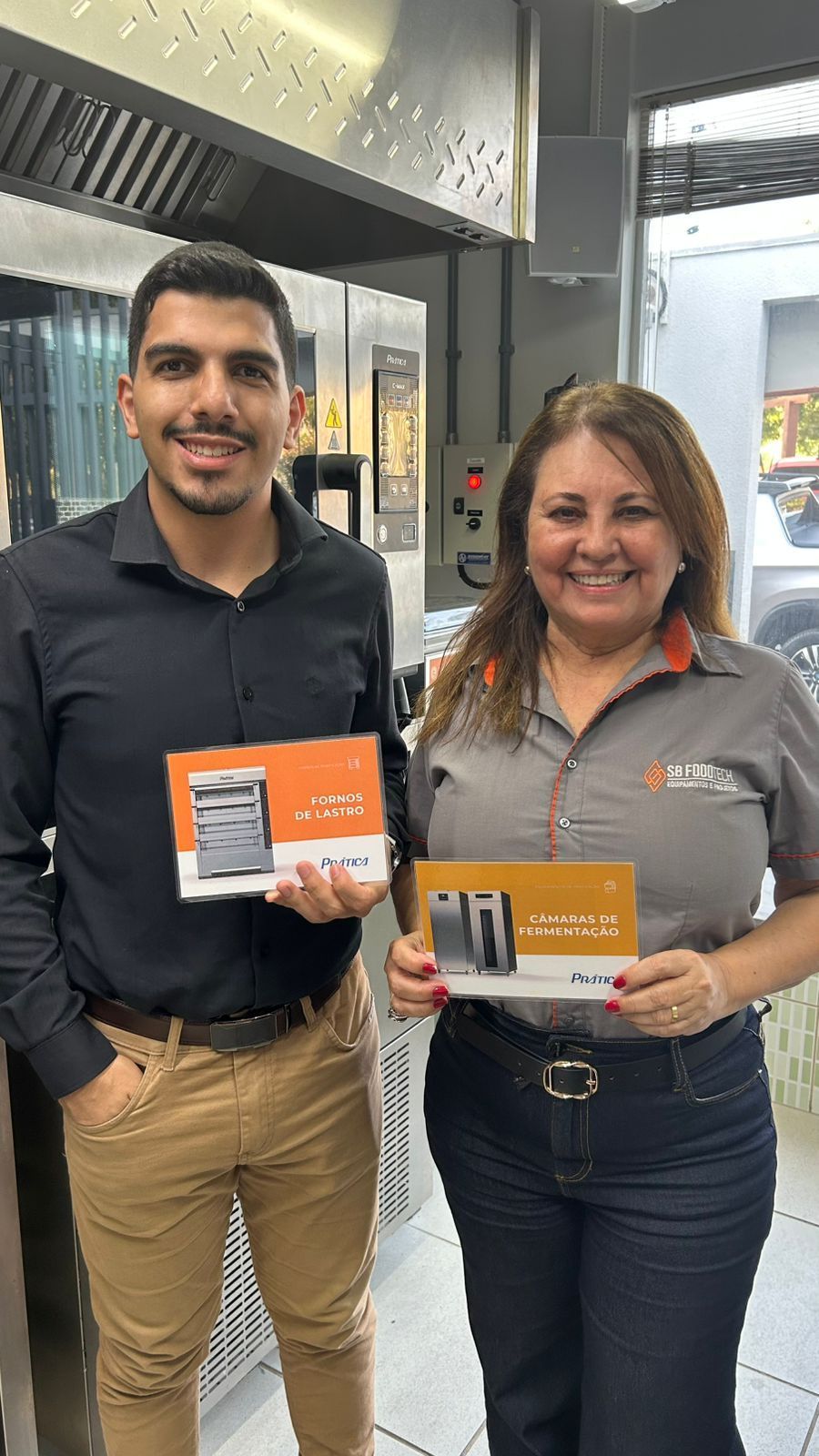Homem e mulher segurando folhetos, sorrindo em frente ao forno, possivelmente de uma padaria.