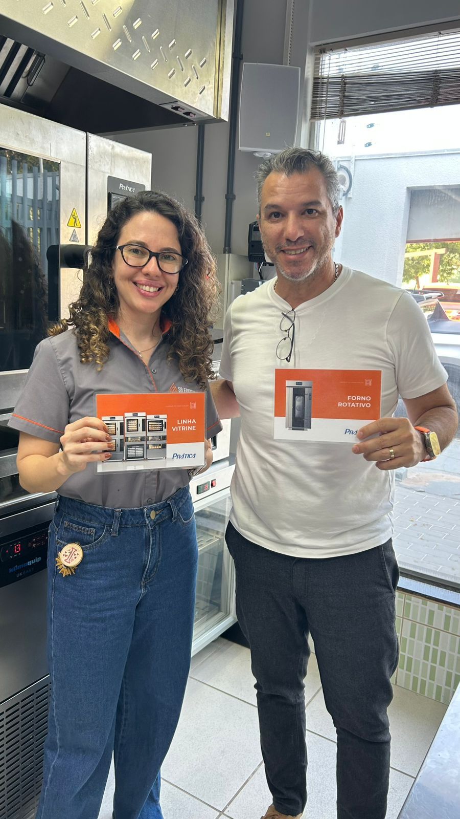 Mulher e homem sorrindo, segurando folhetos em uma loja. Mulher usa óculos e calça jeans. Homem usa camisa branca e óculos.