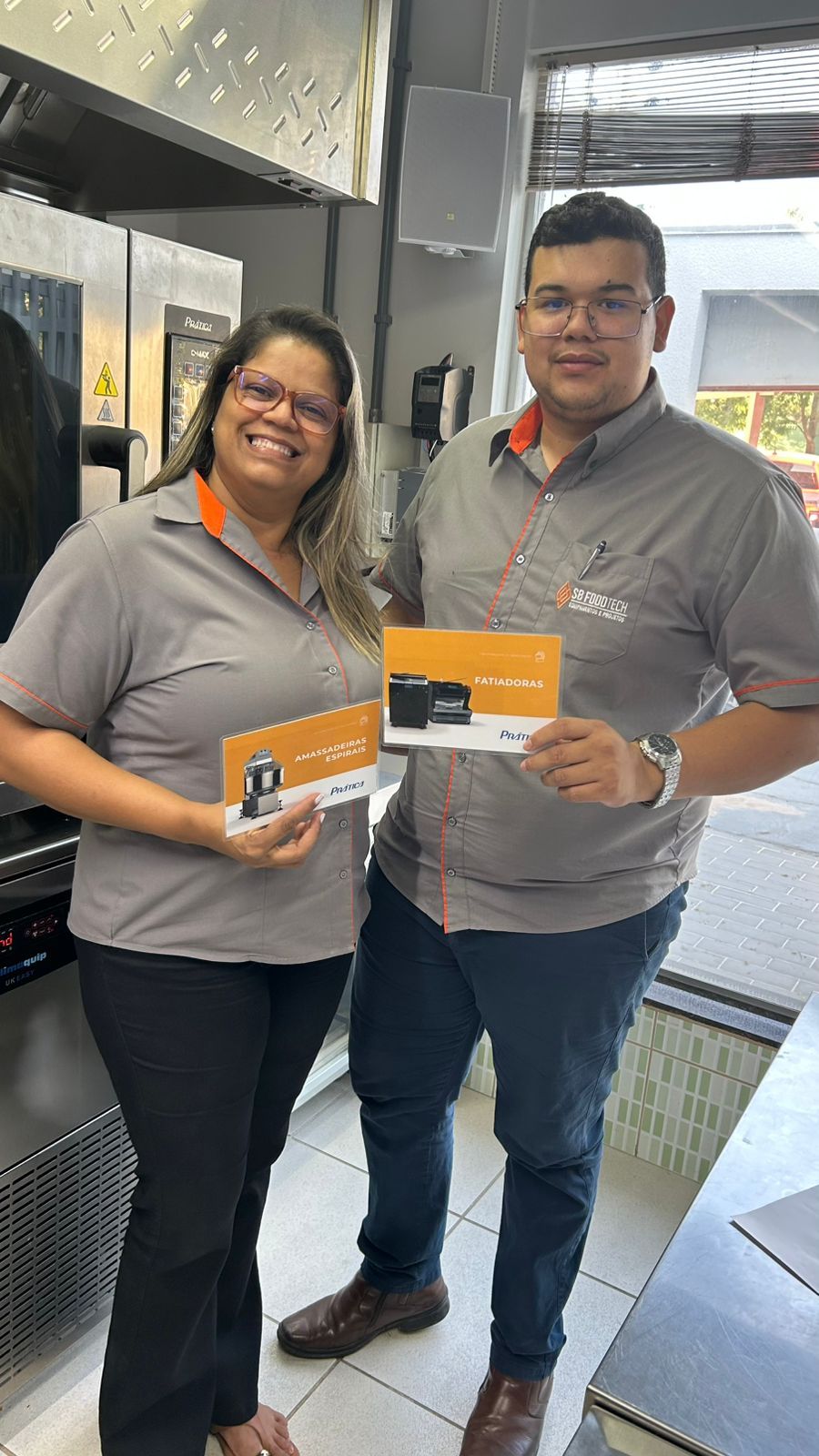 Duas pessoas sorrindo e segurando cartões, vestindo camisas cinza com detalhes em laranja, em uma cozinha.