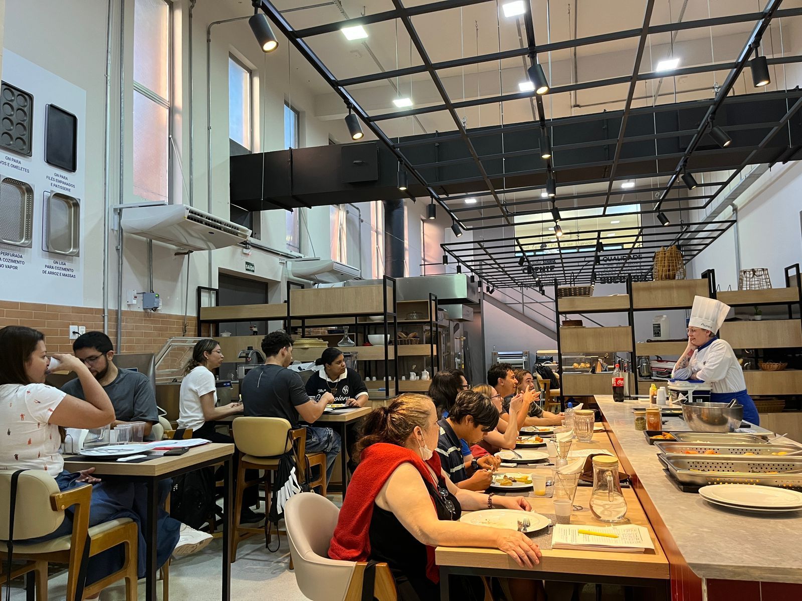 Interior do restaurante: clientes comendo nas mesas; chef de chapéu fala com clientes no balcão.