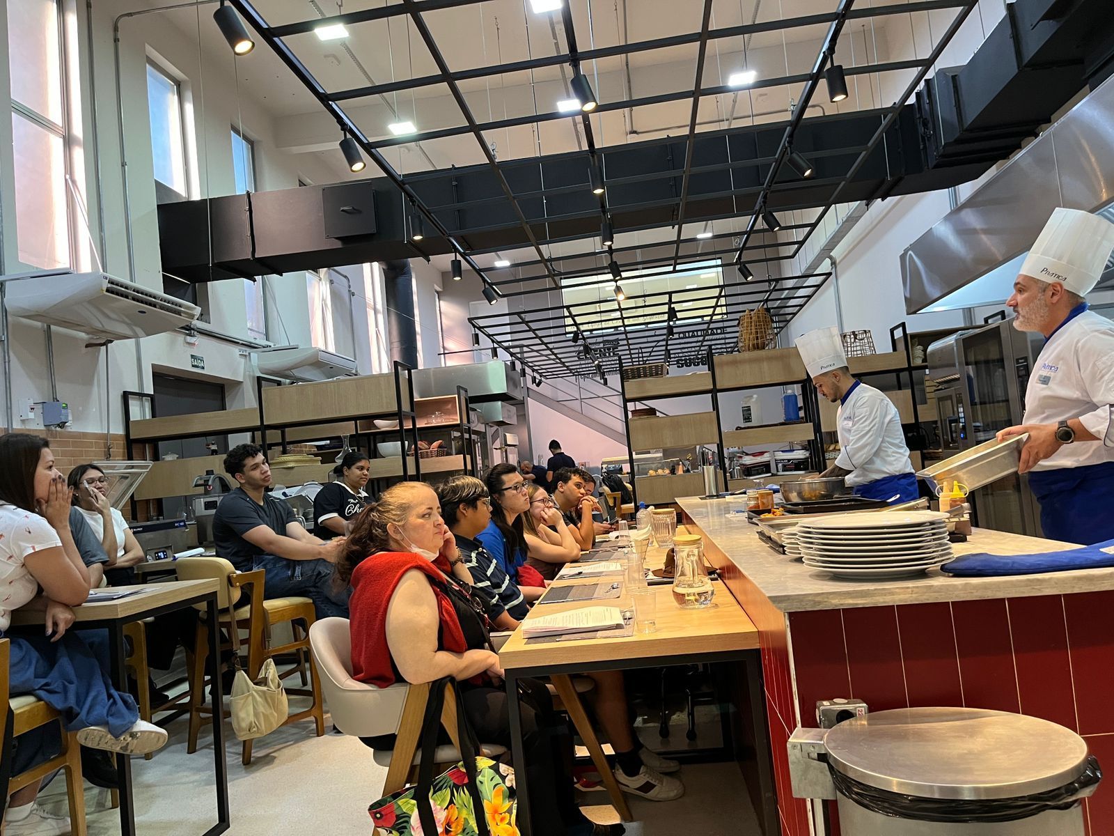 Chef demonstrando culinária para um grupo em uma cozinha. Pessoas sentadas às mesas observam atentamente.