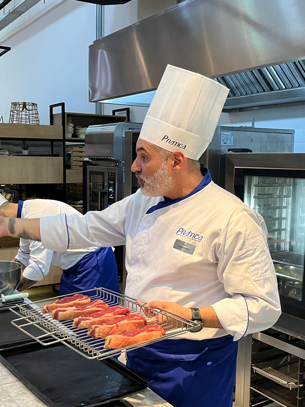 Chef de uniforme branco, segurando carnes grelhadas e gesticulando em uma cozinha profissional.