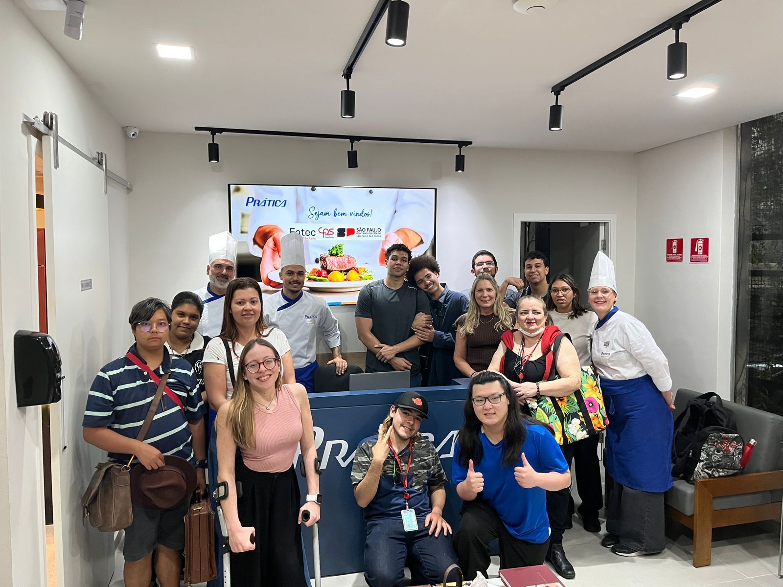Grupo de pessoas sorrindo em uma cozinha. Algumas usam chapéus de chef, em pé perto de uma tela com comida.