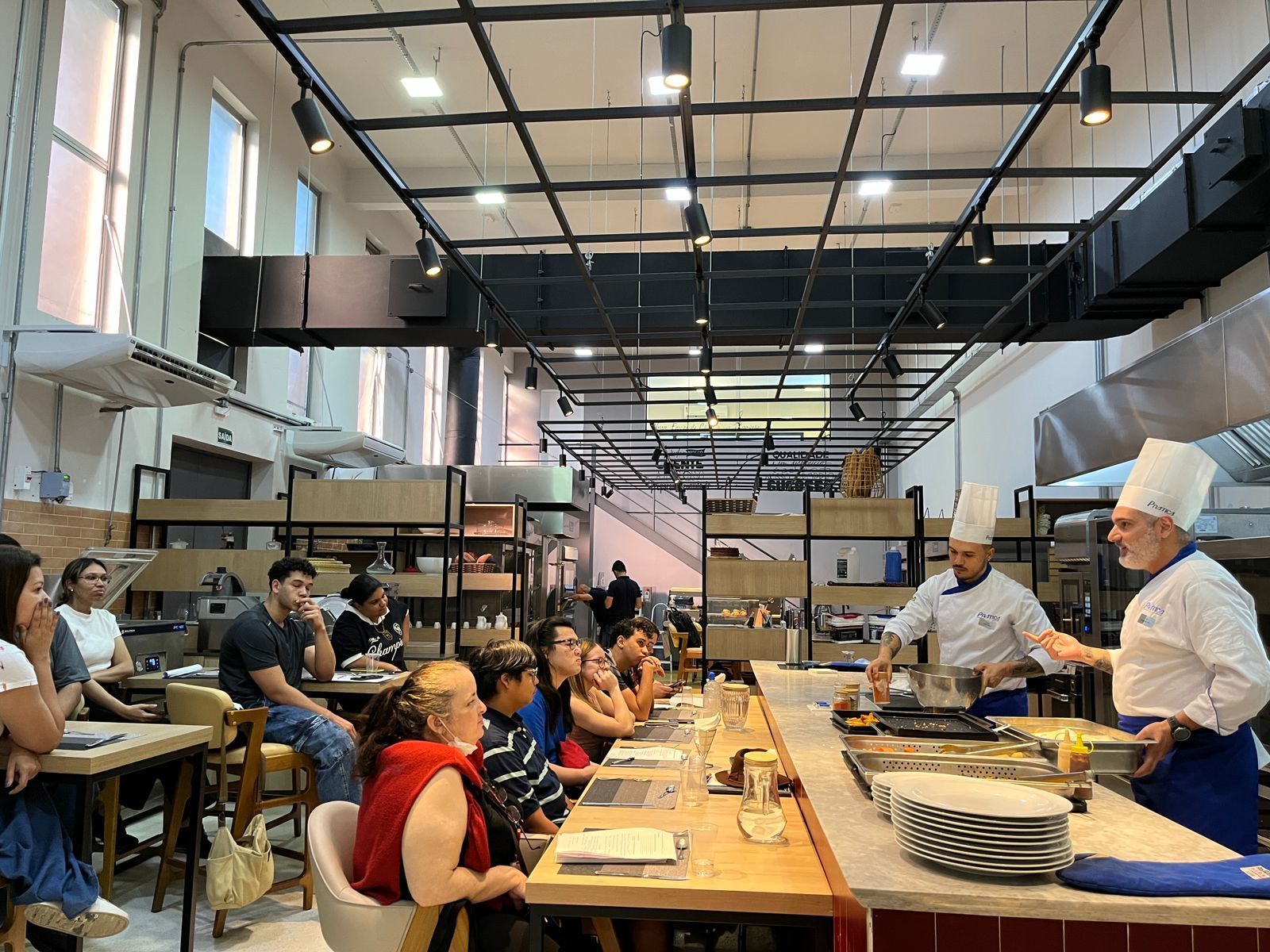 Chef demonstrando culinária para uma turma em uma cozinha grande. Alunos observam.