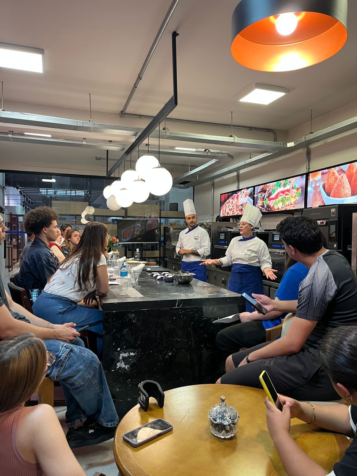 Chefs demonstram culinária para um grupo de pessoas em torno de uma ilha na cozinha. Dentro de um restaurante.