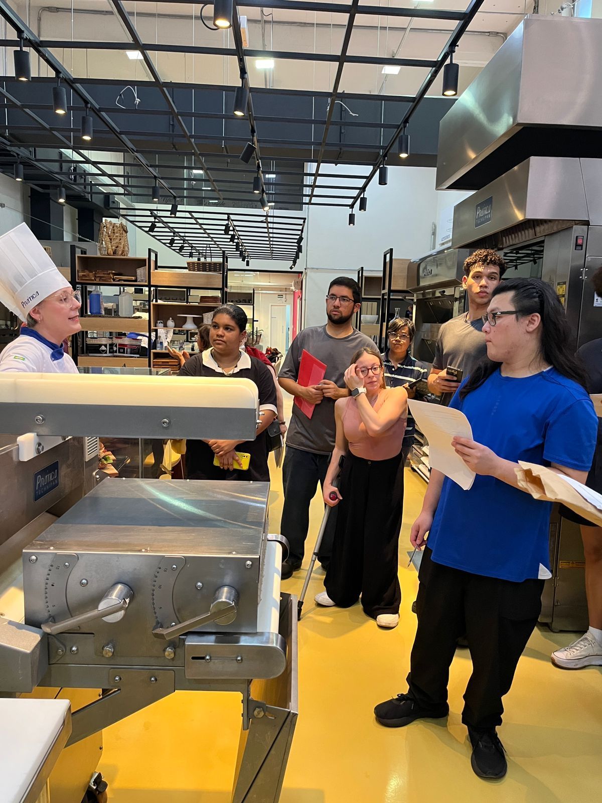 Chef instruindo alunos em uma cozinha comercial, alunos observam e ouvem.