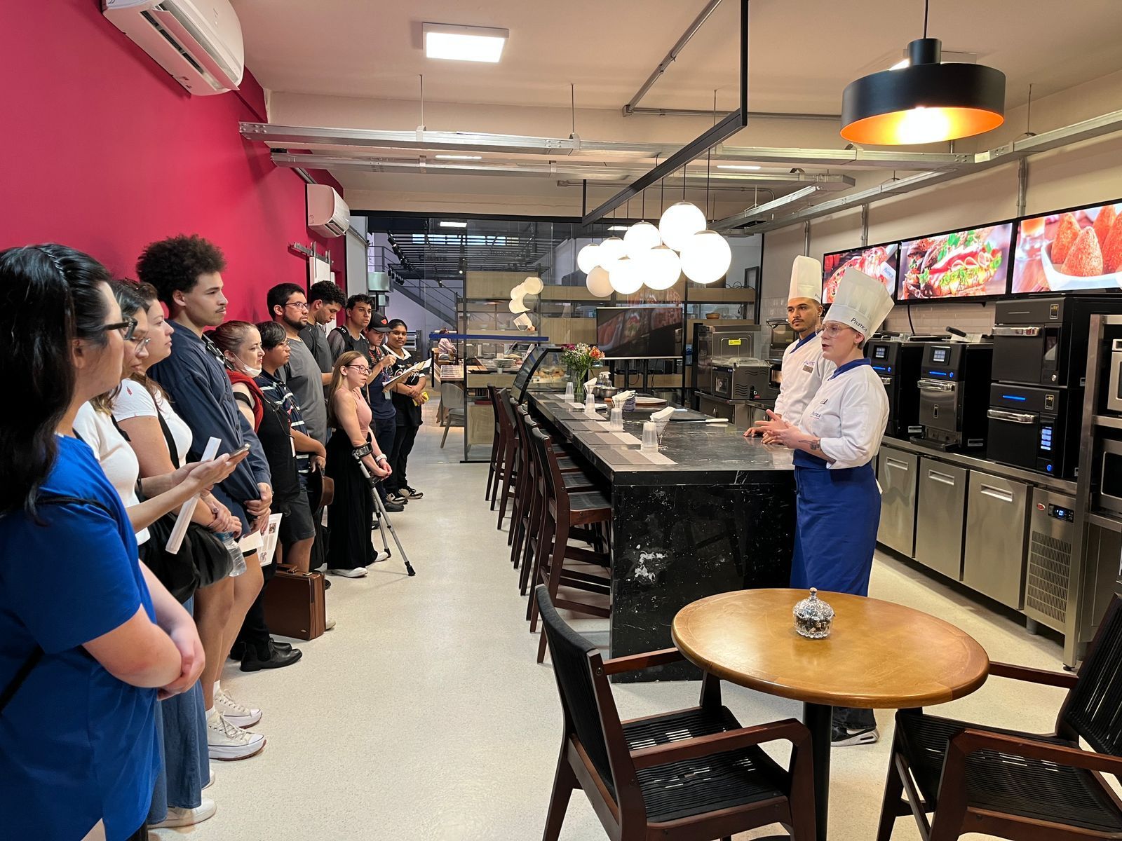 Um chef fala para um grupo em uma cozinha. As pessoas ficam em pé, ouvindo, perto de um balcão e mesas.