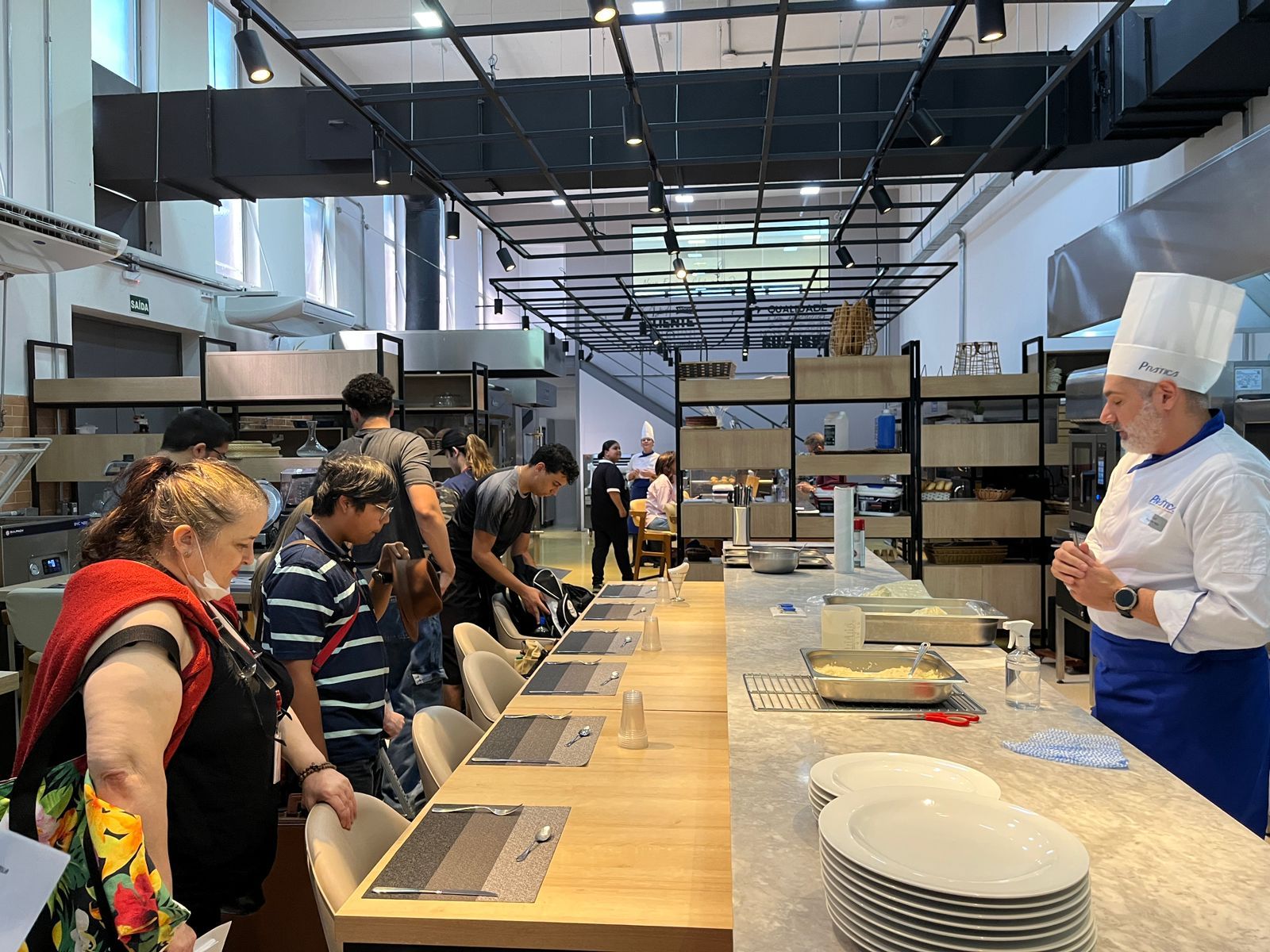 Chef dando instruções em uma aula de culinária. As pessoas observam em um balcão comprido com pratos.
