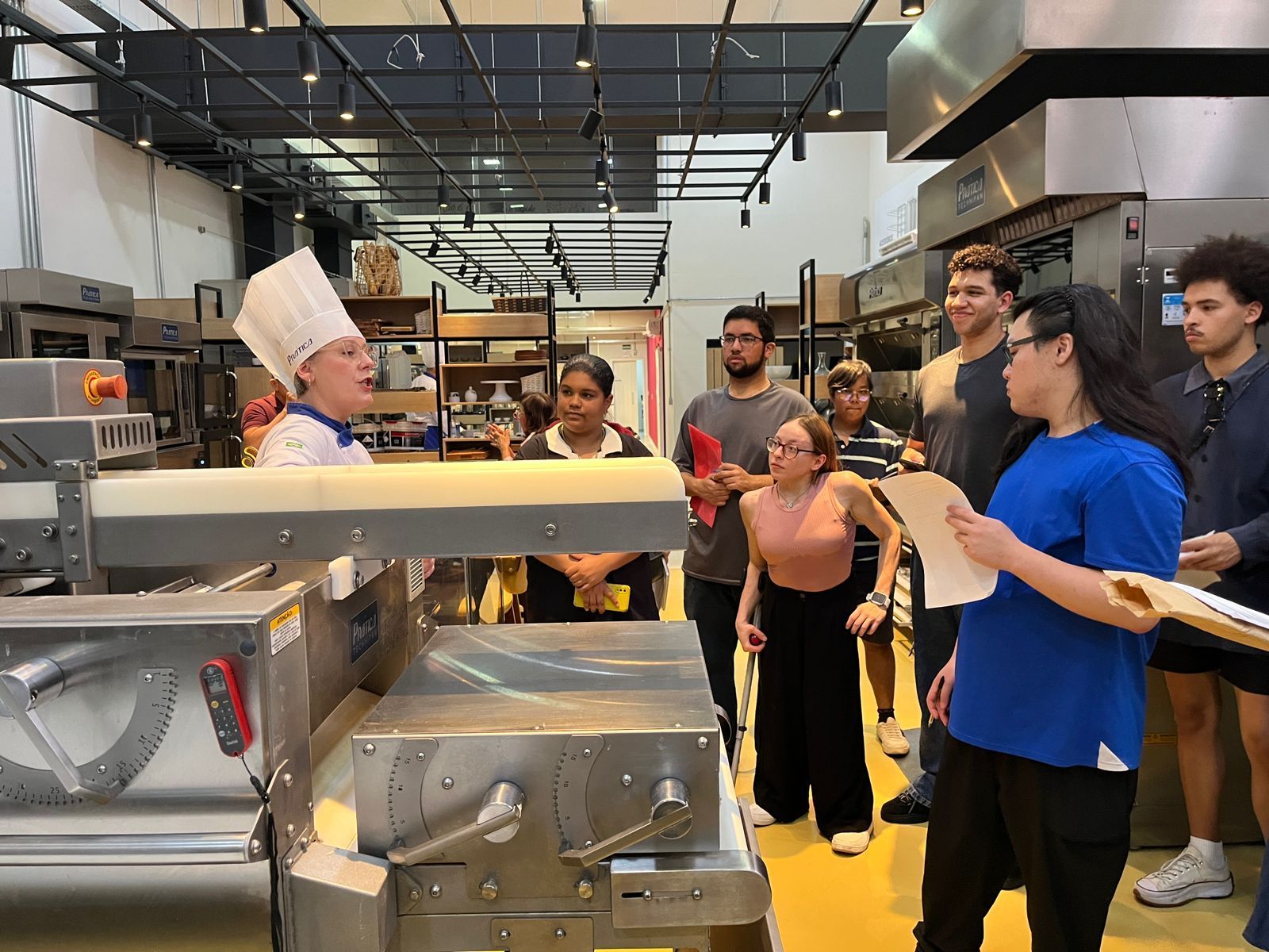 Chef de chapéu branco instrui um grupo na cozinha; equipamentos de aço inoxidável visíveis, alunos segurando papéis.