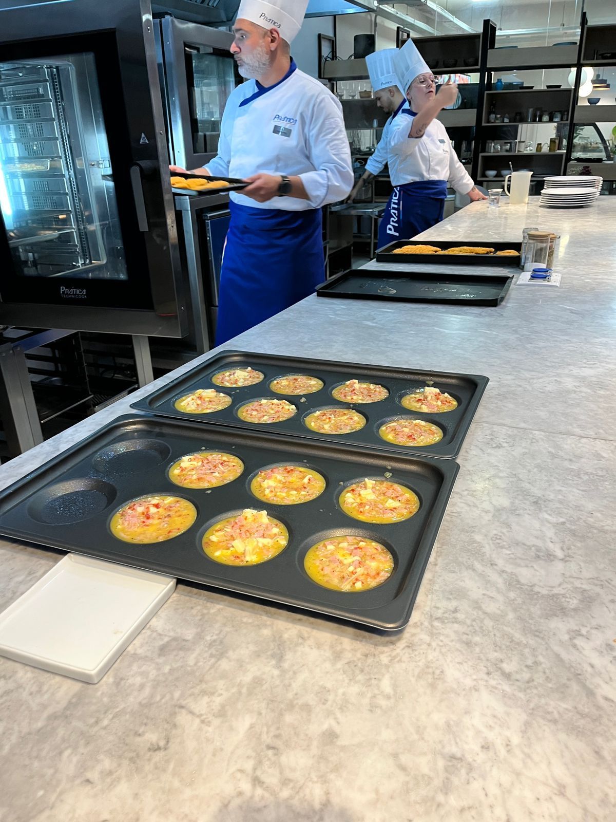 Chefs preparando comida na cozinha, com produtos assados em bandejas no balcão.