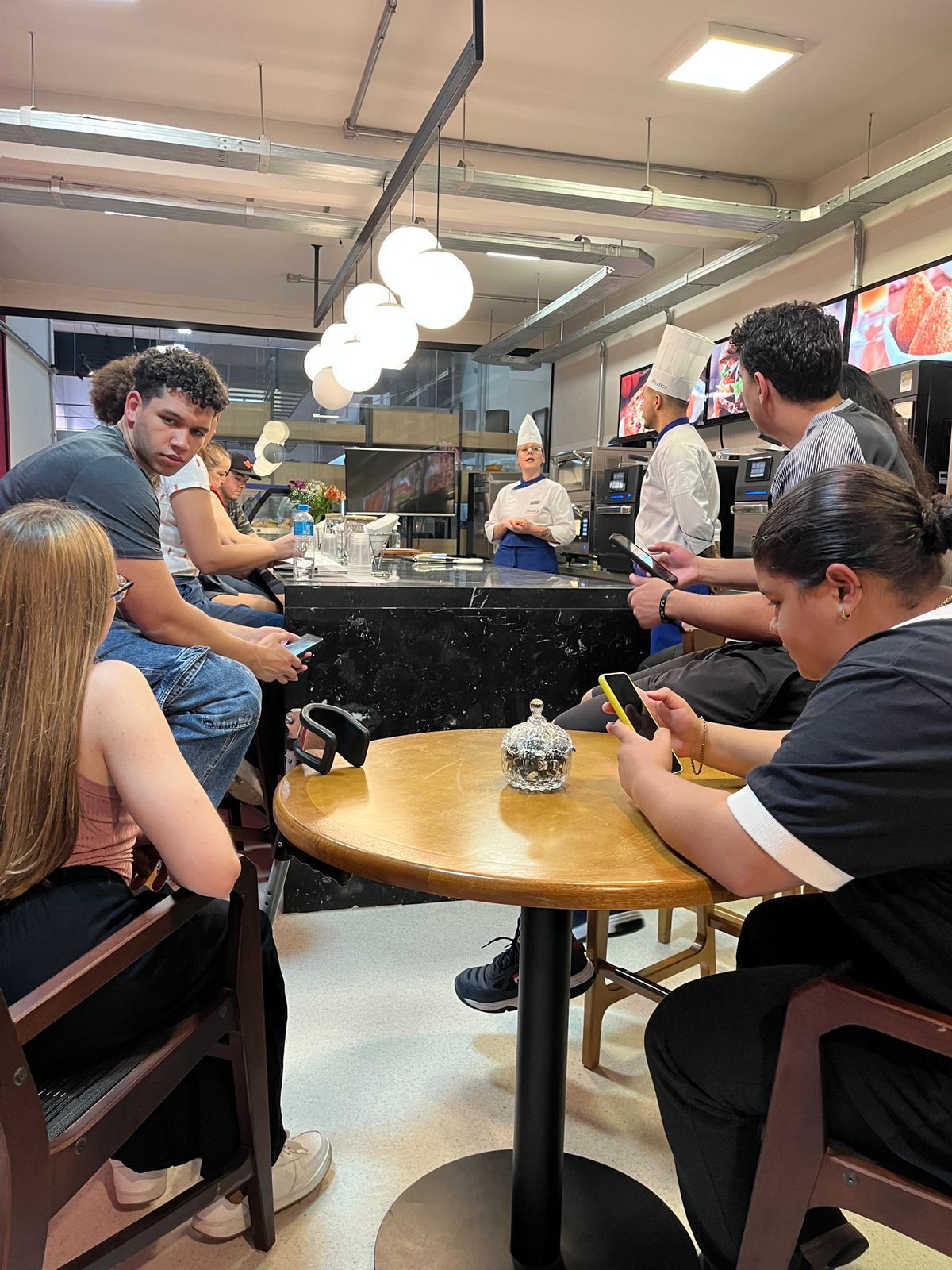 Pessoas no balcão do café. Chefs de branco, clientes nas mesas, checando celulares. Cores neutras.