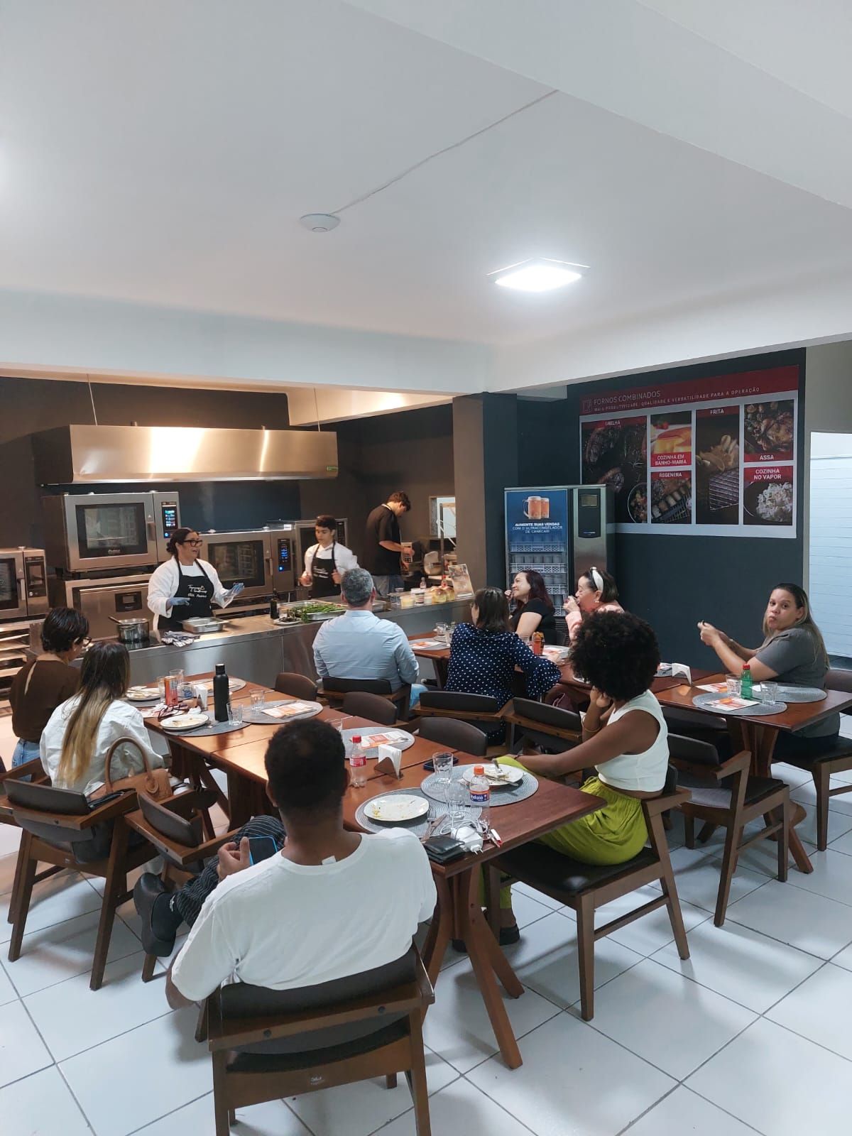 As pessoas observam os chefs na cozinha, sentados às mesas em uma área de jantar; espaço de restaurante moderno com demonstração de culinária.