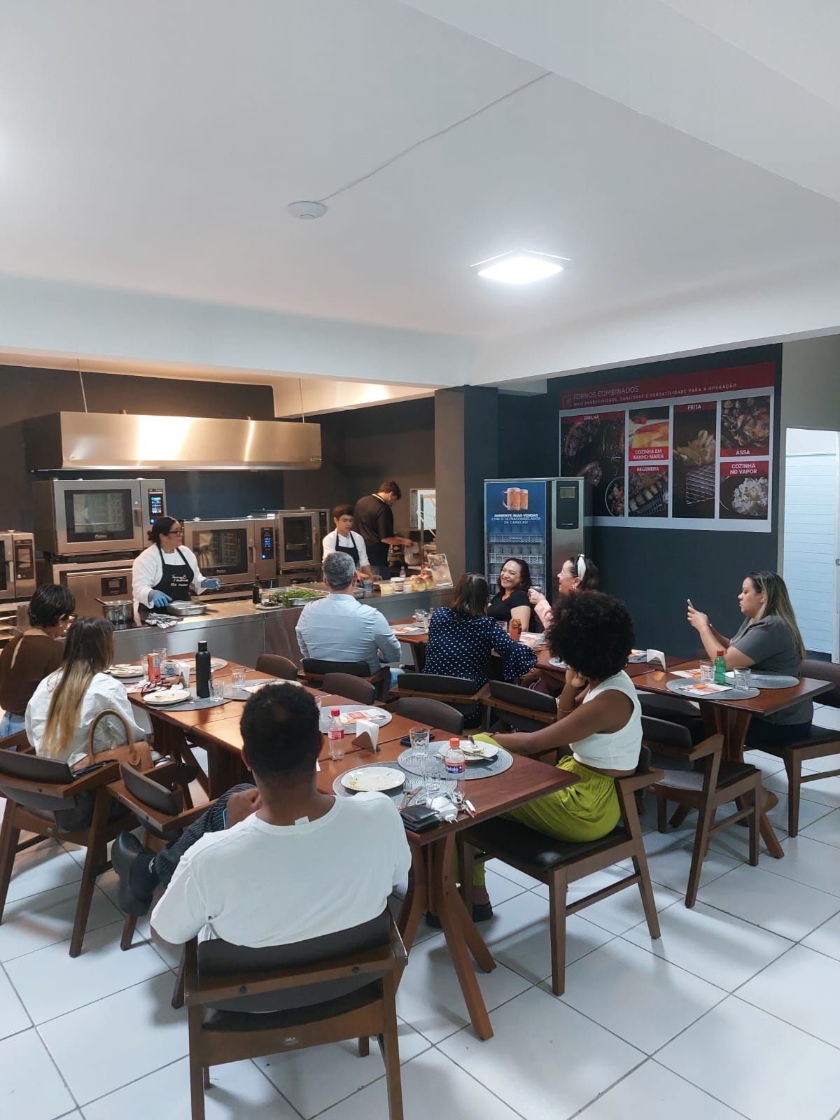 Pessoas sentadas às mesas, observando chefs em uma cozinha profissional, provavelmente uma aula de culinária ou demonstração.