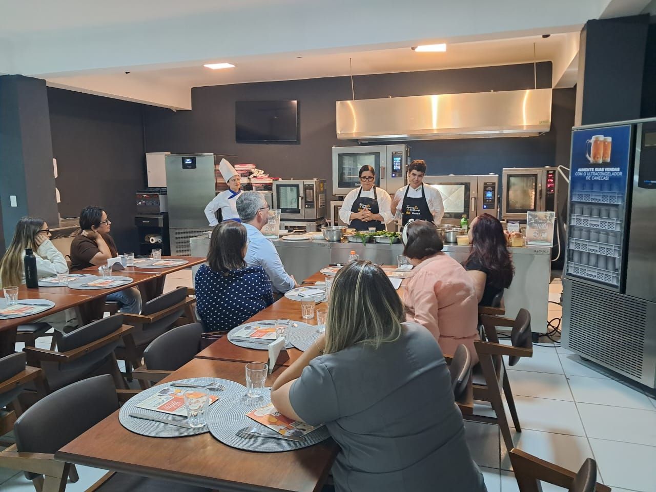 Uma demonstração de culinária com dois chefs preparando comida e um grupo de pessoas assistindo nas mesas de um restaurante.