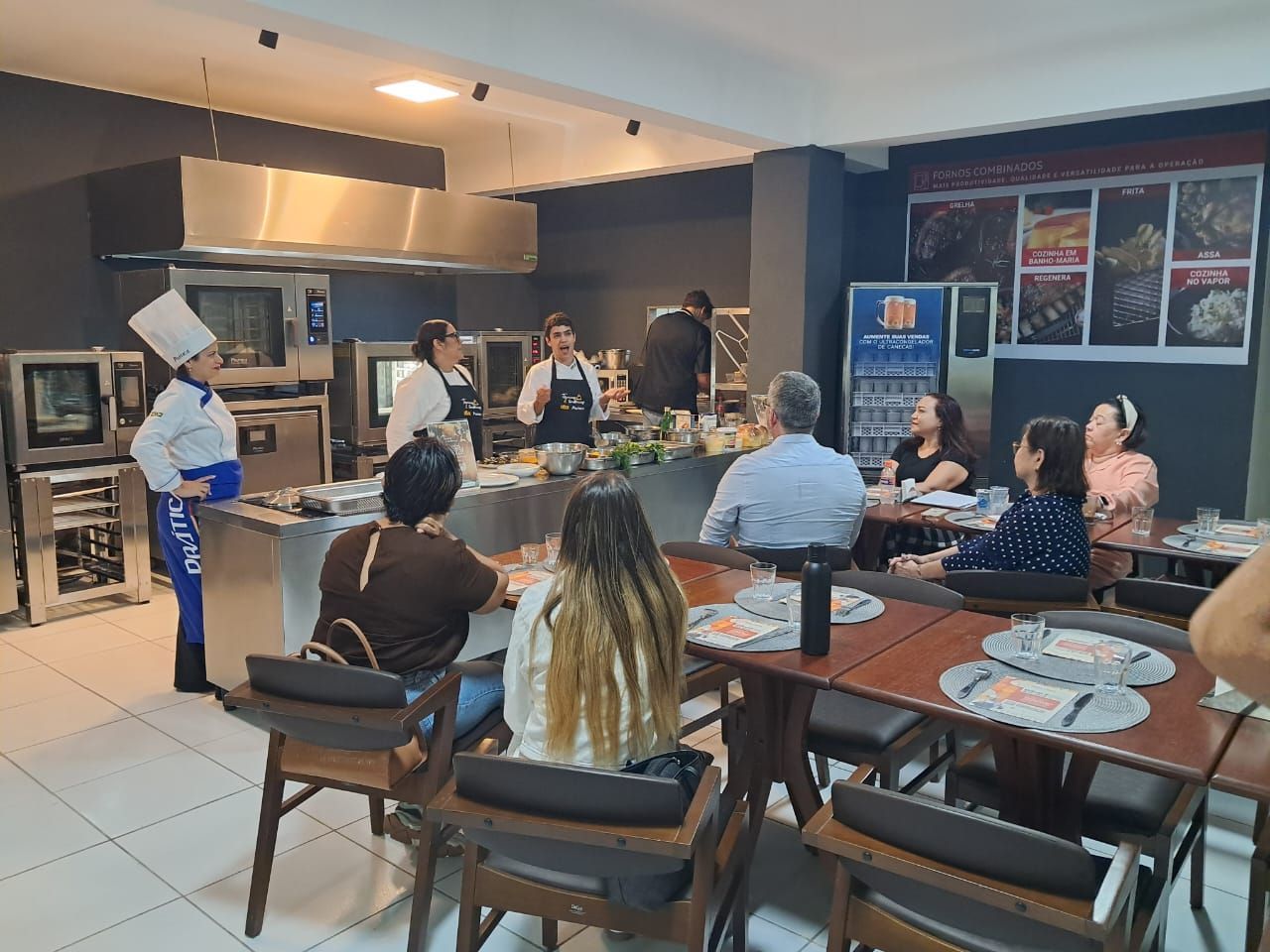 Aula de culinária: Chef dando instruções na cozinha. Participantes observam nas mesas.