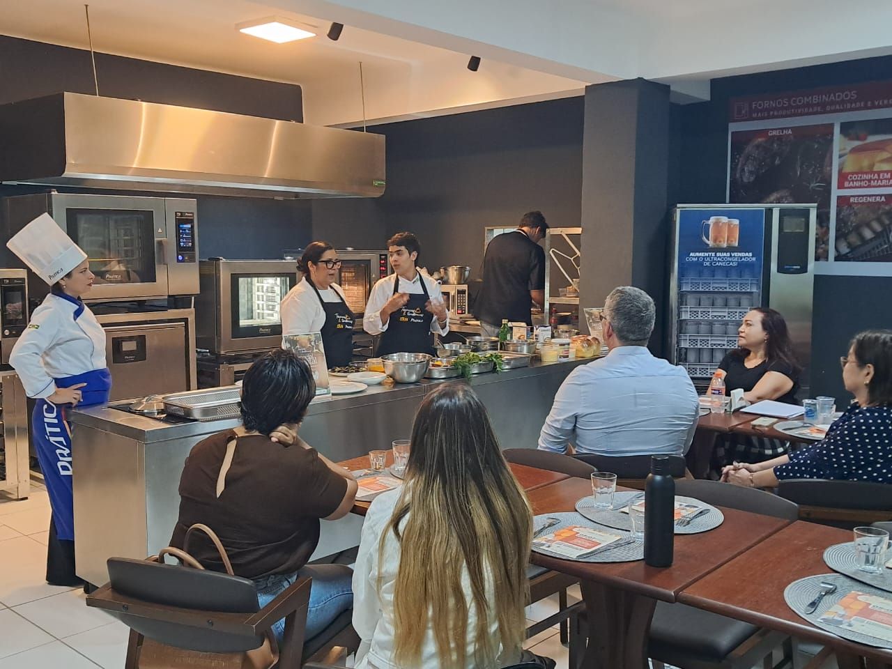 Chef e cozinheiros demonstrando uma receita para um pequeno público em uma cozinha moderna.