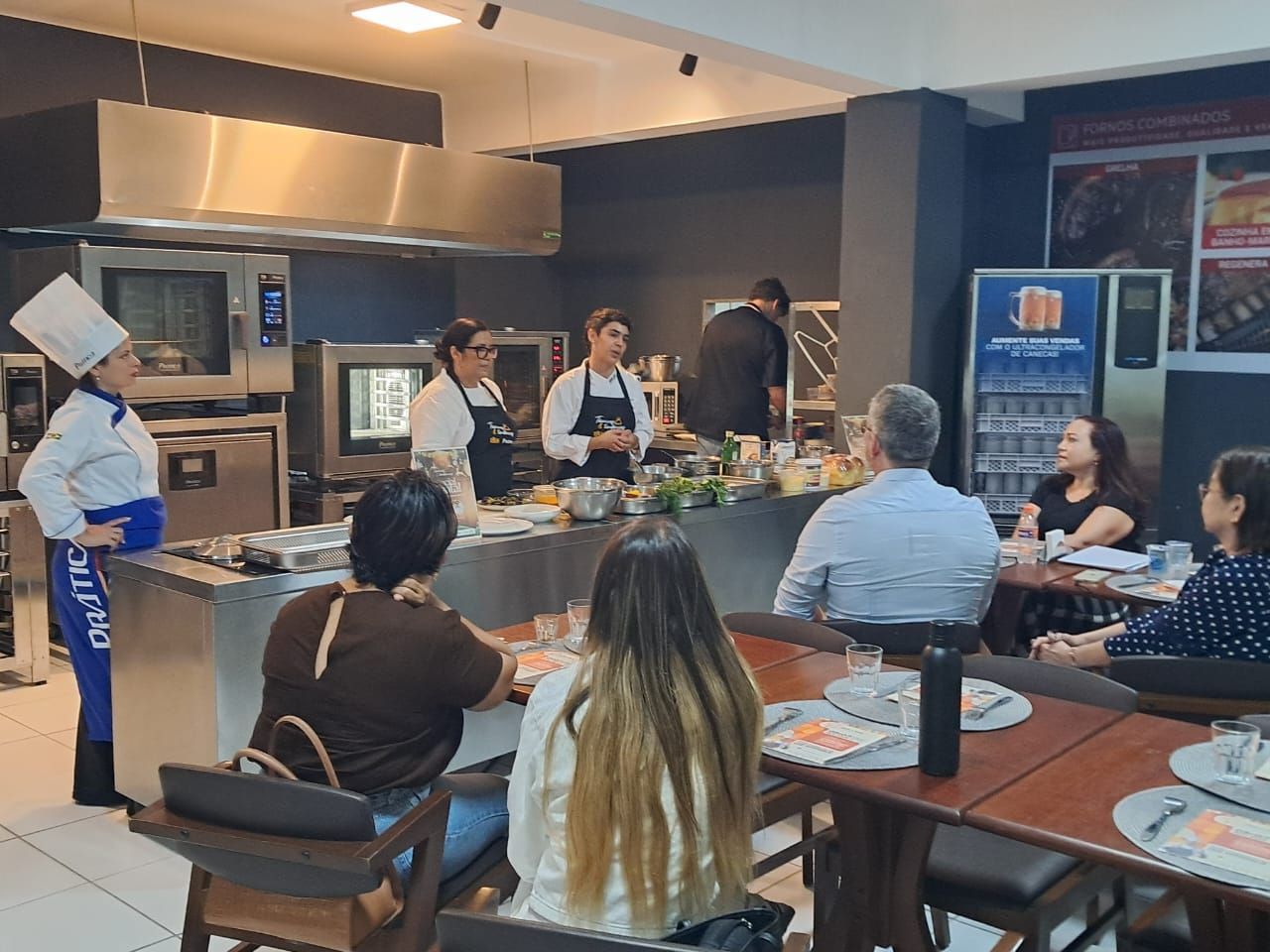 Chef demonstra culinária em uma cozinha para um público sentado em mesas.