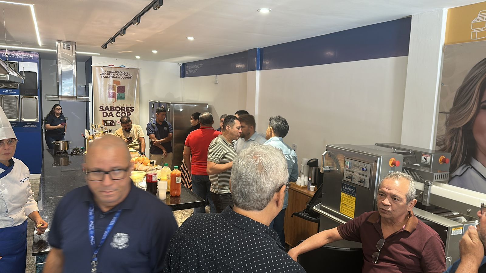 Uma demonstração em uma cozinha lotada. Chefs e público observam os equipamentos e a preparação dos pratos.
