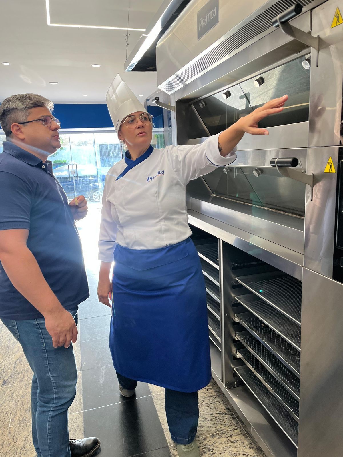 Um chef uniformizado aponta para um forno, explicando-o a um homem vestido casualmente.