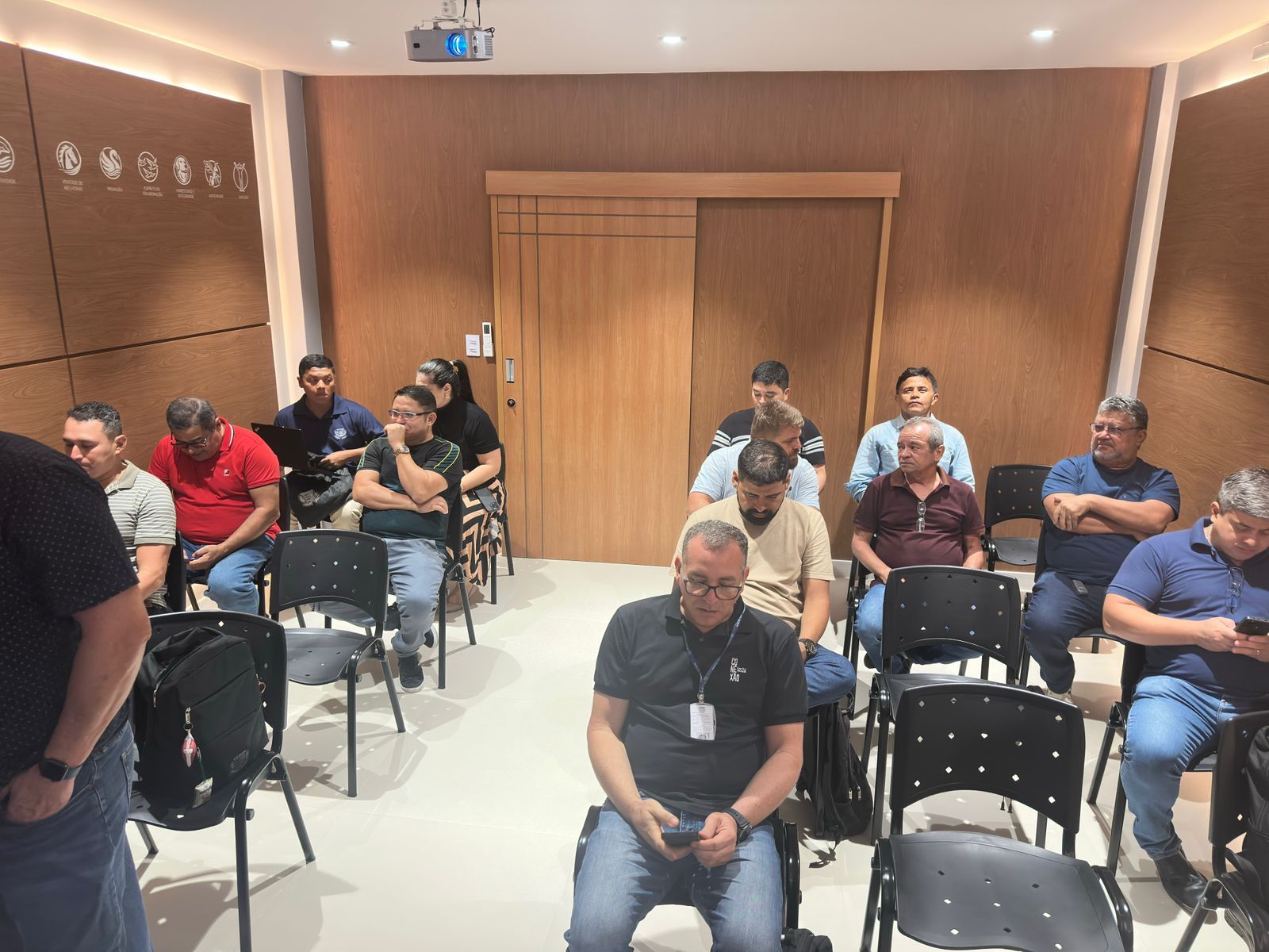 Pessoas sentadas em uma sala de conferências, ouvindo. Paredes de madeira e uma tela de projeção são visíveis.