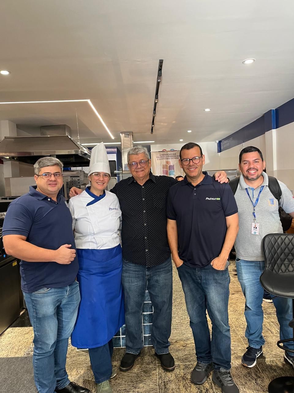 Cinco pessoas sorrindo; um chef e homens em roupas casuais, em ambientes fechados, posando.