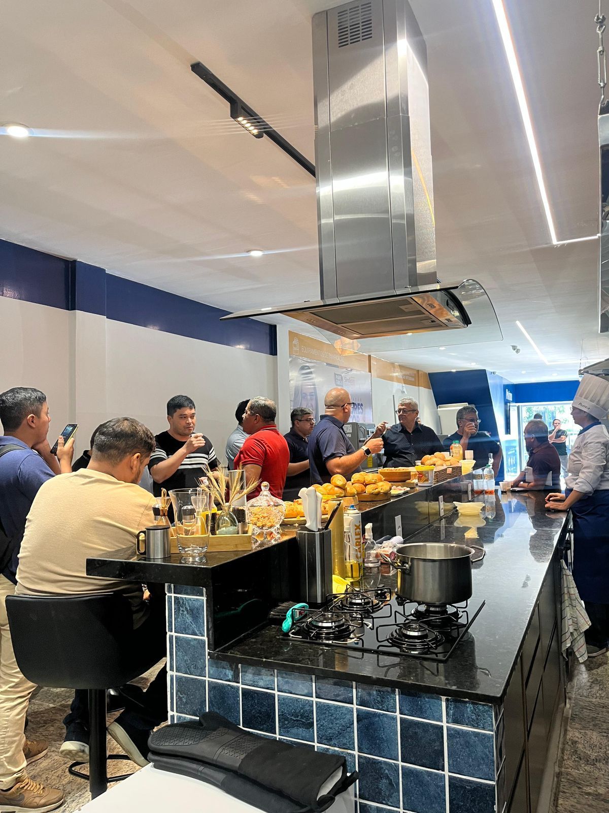 Pessoas em um bar; comida sendo servida. Balcão longo, azulejos azuis, ventilação de aço inoxidável.