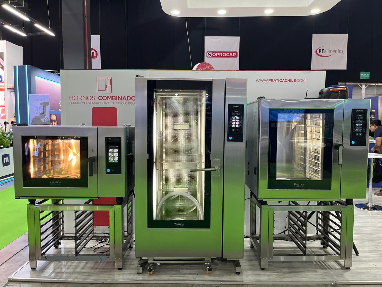 Três fornos comerciais de aço inoxidável em exposição em uma feira. Paredes verdes, luzes brilhantes.