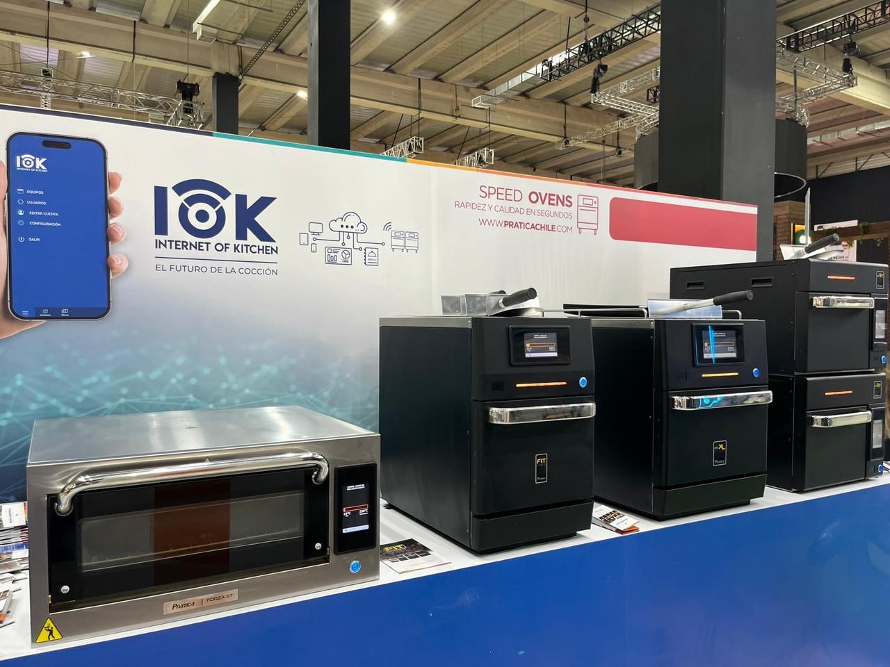 Um estande de feira comercial exibindo várias máquinas de serviço de alimentação pretas e um pequeno forno. Um celular exibe uma interface de aplicativo.