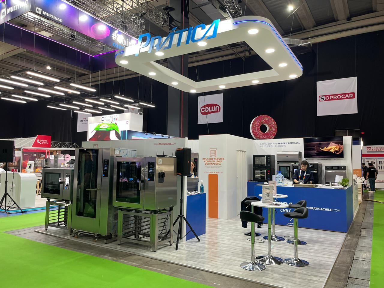 Estande de feira com letreiro iluminado. Máquinas em exposição, pessoas e um piso verde brilhante.