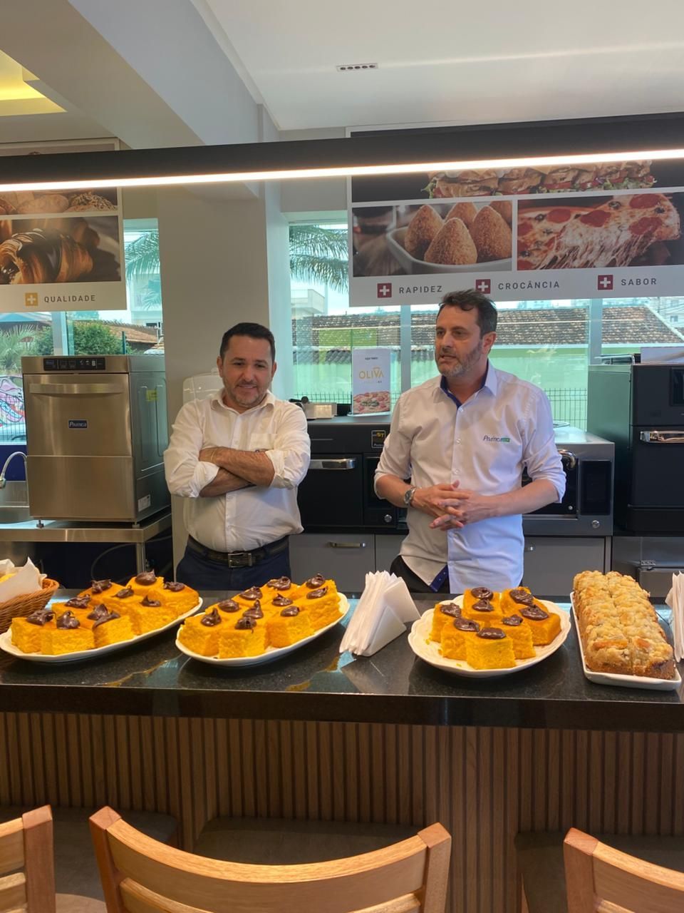 Dois homens atrás de um balcão com comida; bolos e doces.