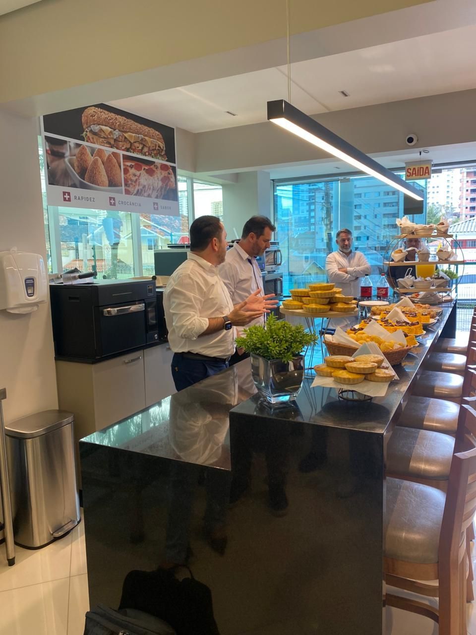 Pessoas no balcão de um café; três homens de camisas brancas, exposição de alimentos, ambiente moderno.