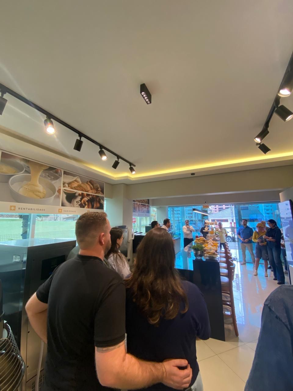 Pessoas em uma padaria bem iluminada, olhando para um balcão com comida; vitrine de vidro ao fundo.