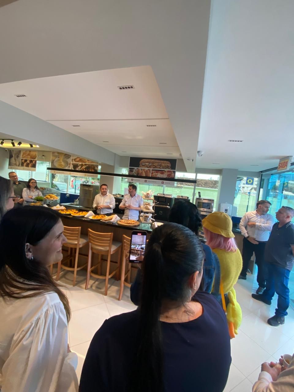 Pessoas assistem à demonstração de um chef no balcão de um restaurante; comida visível e vista externa.