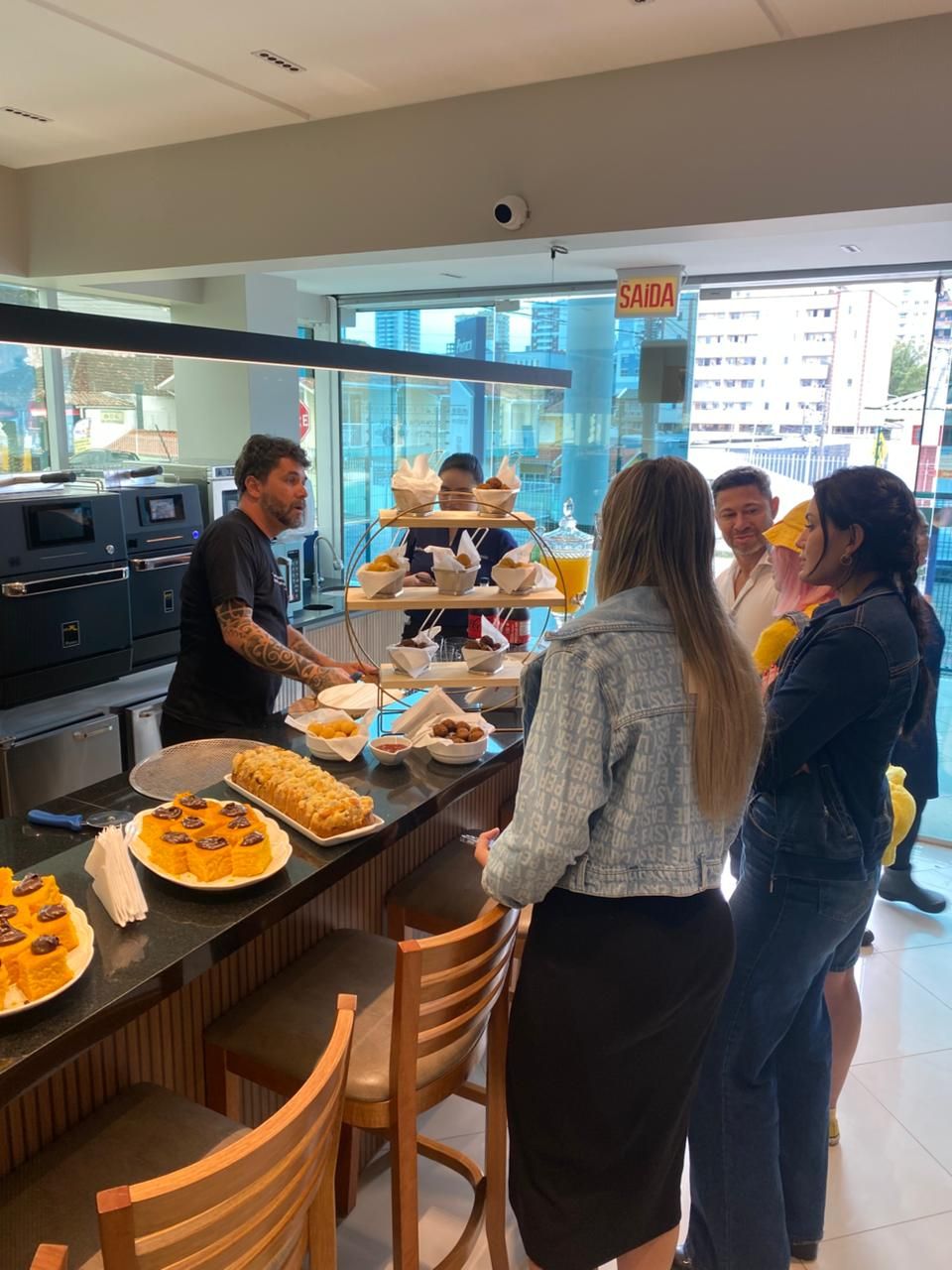 Um homem prepara comida no balcão de um café, interagindo com vários clientes.