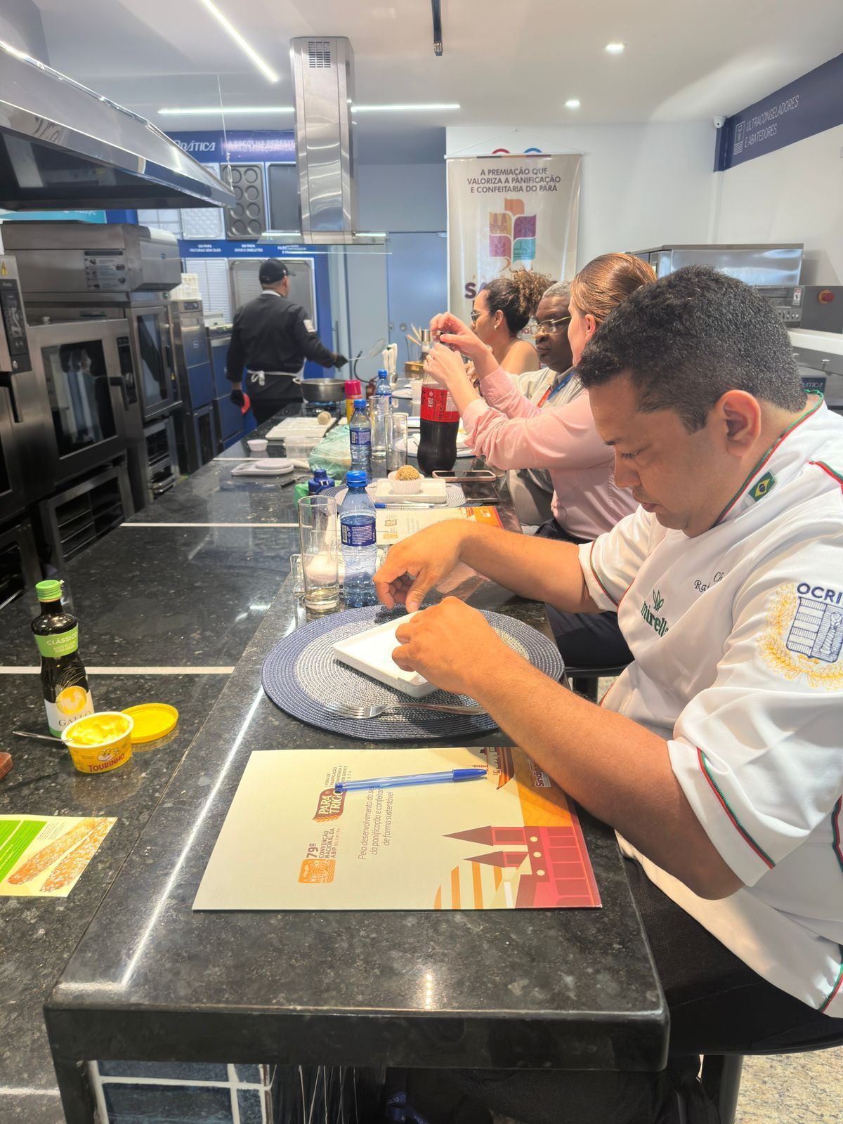 Chef de uniforme branco preparando comida no balcão da cozinha. Outros trabalham atrás dele.