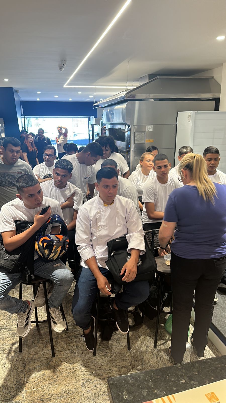 Chefs de jaleco branco sentados e em pé ouvindo uma mulher em uma cozinha ou restaurante.