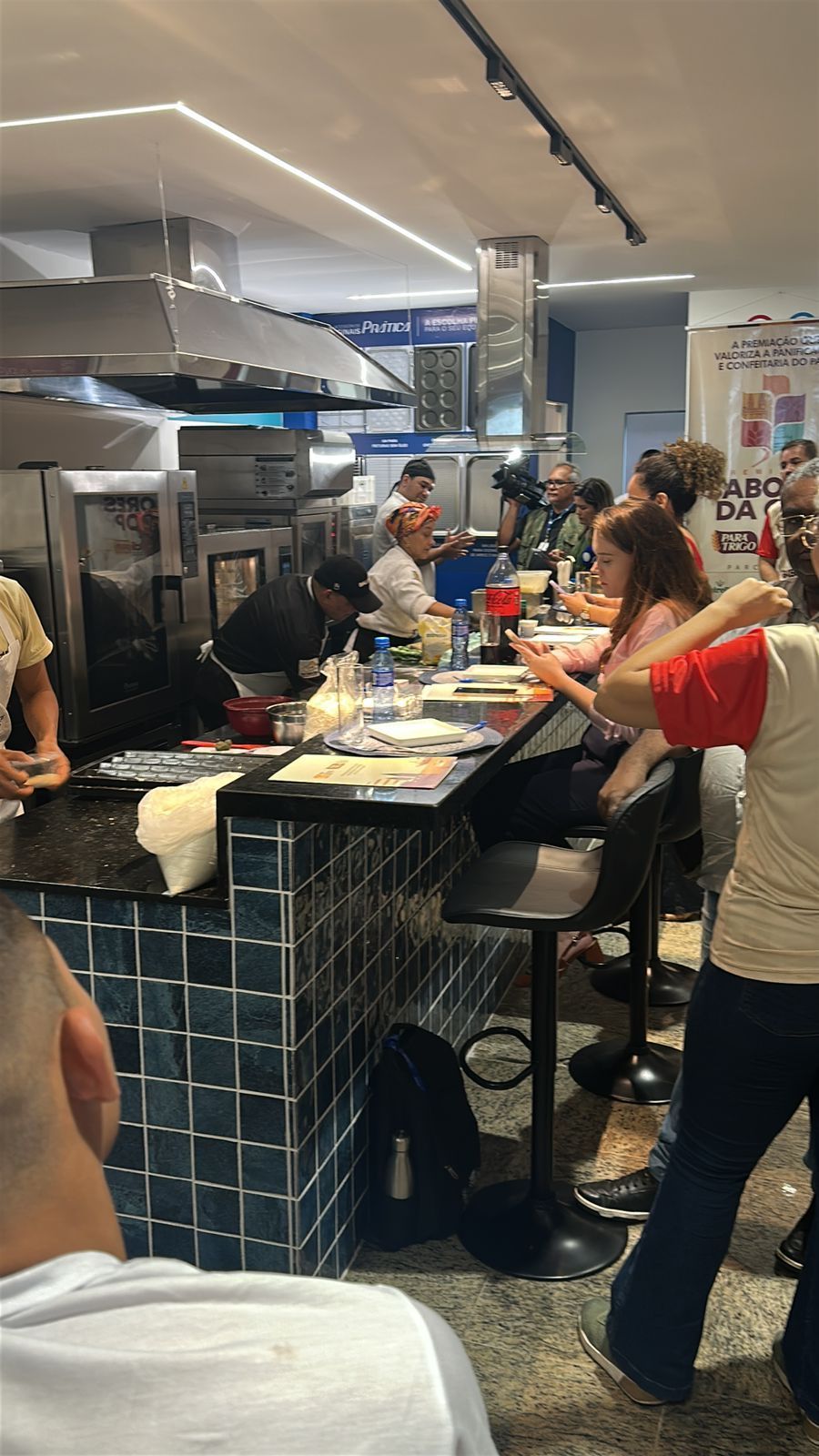 Balcão de restaurante com pessoas comendo, chef trabalhando; balcão com azulejos pretos e azuis, eletrodomésticos de aço inoxidável.