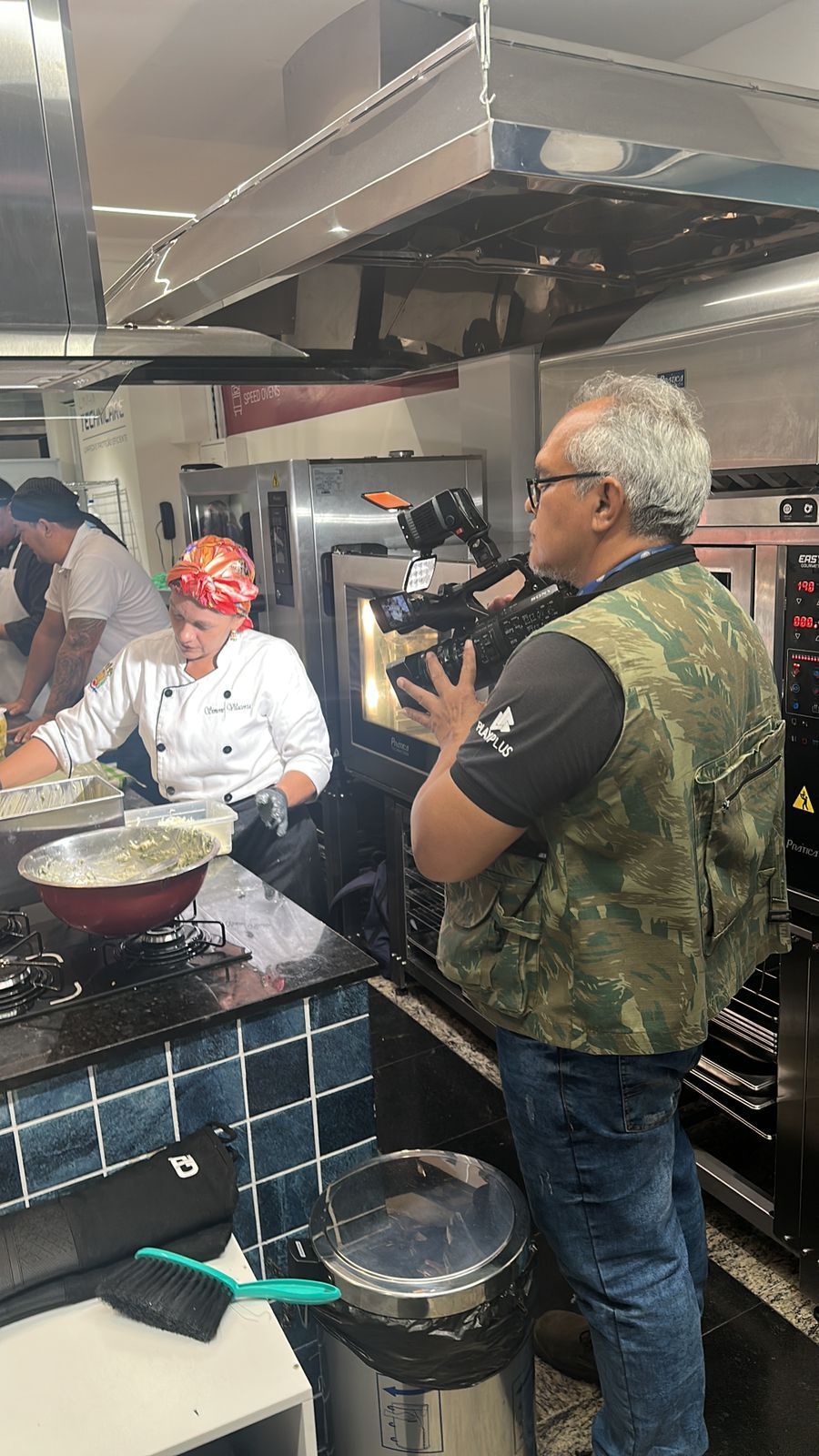 Uma pessoa filma um chef cozinhando em uma cozinha comercial; duas outras pessoas são visíveis.