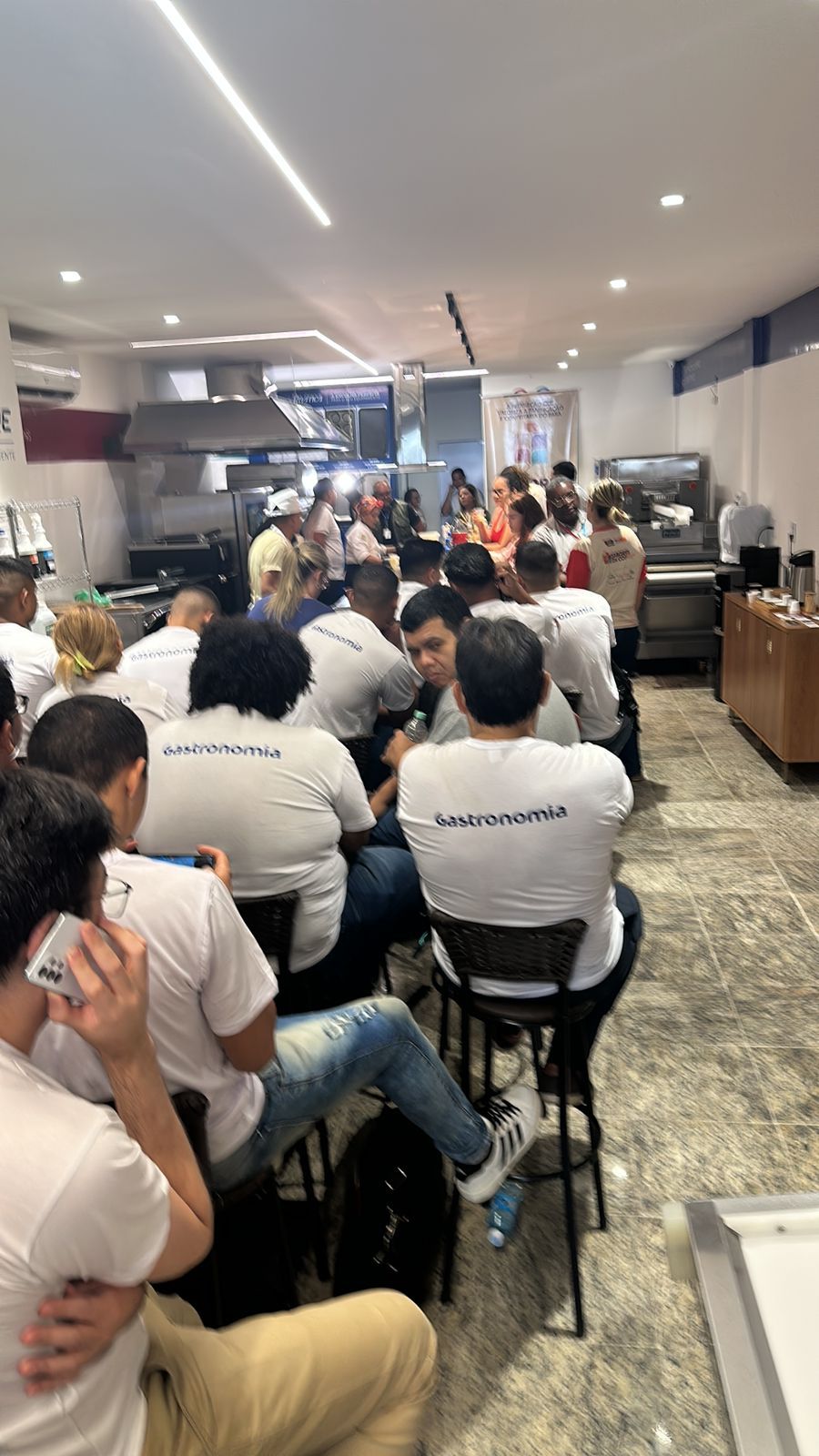 Pessoas de camisa branca participam de uma oficina, viradas para a frente em uma cozinha, algumas usando celulares.