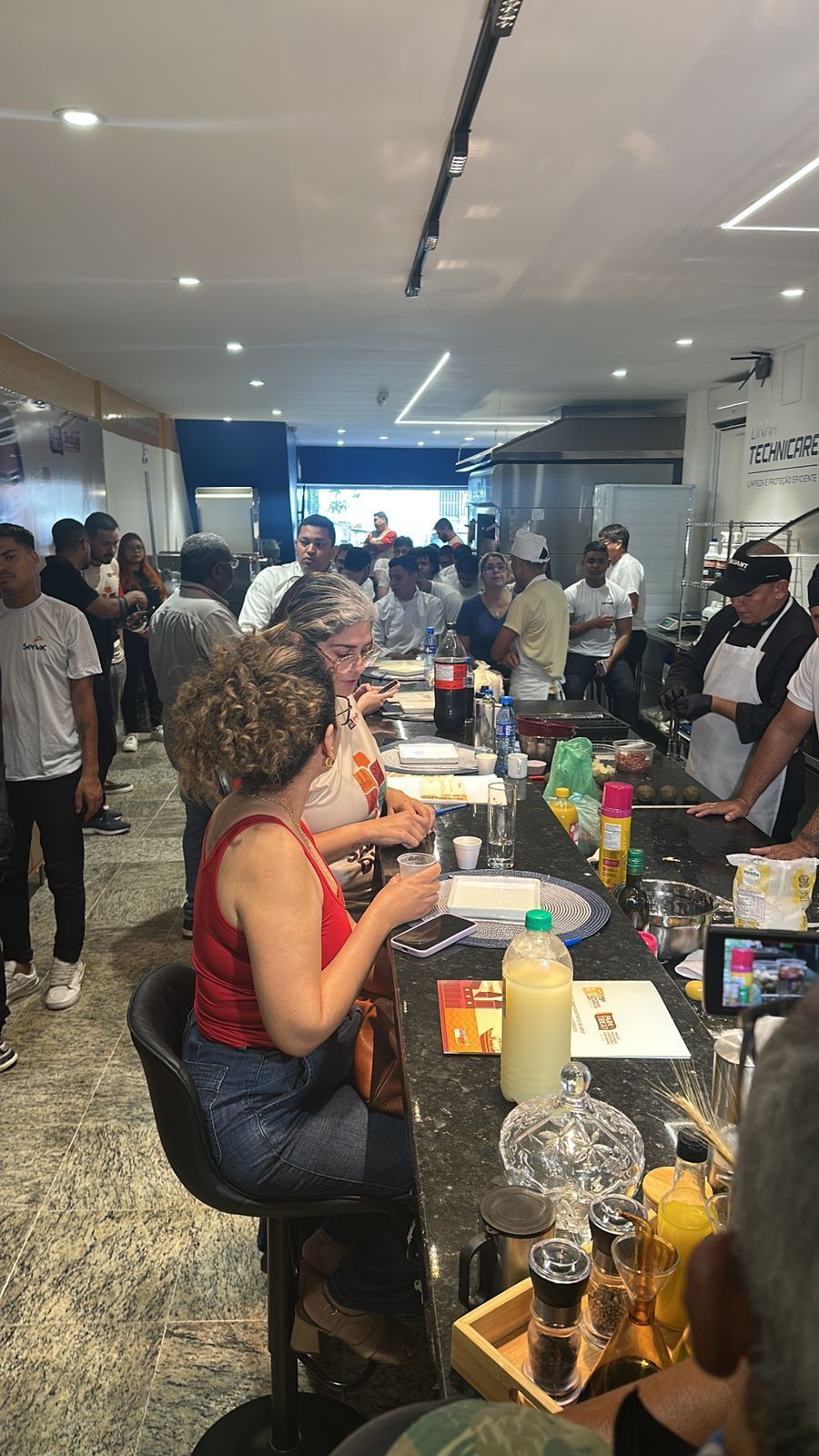 Pessoas no balcão movimentado de um café, algumas sentadas, outras em pé. Garrafas, bebidas e alimentos são visíveis.