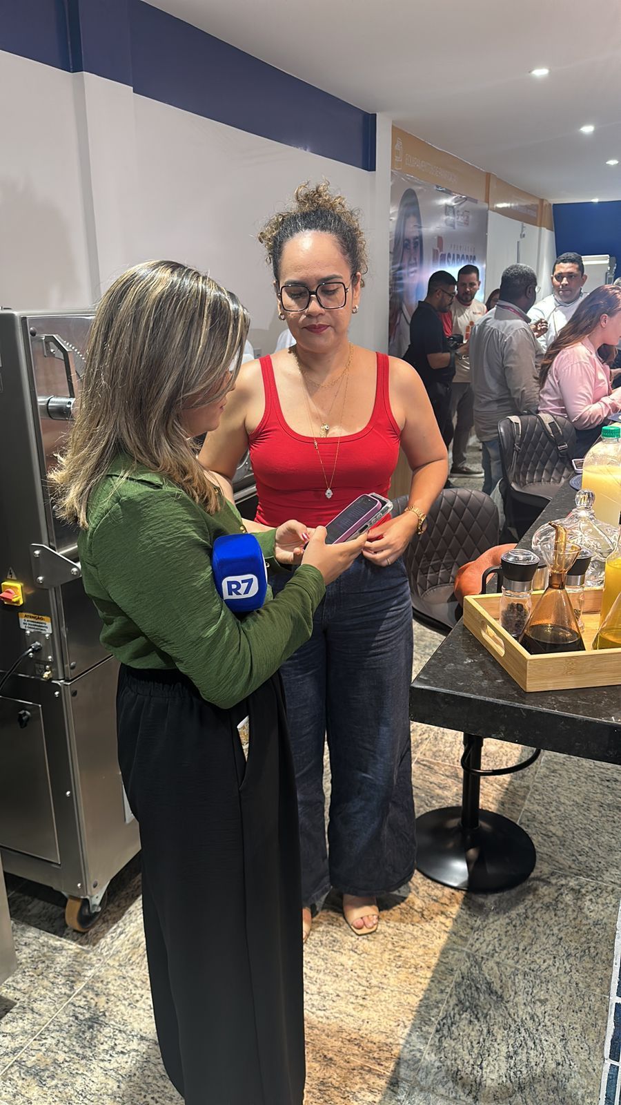 Uma mulher de blusa vermelha é entrevistada. Um repórter de verde segura um telefone. Cenário interno, outras pessoas presentes.