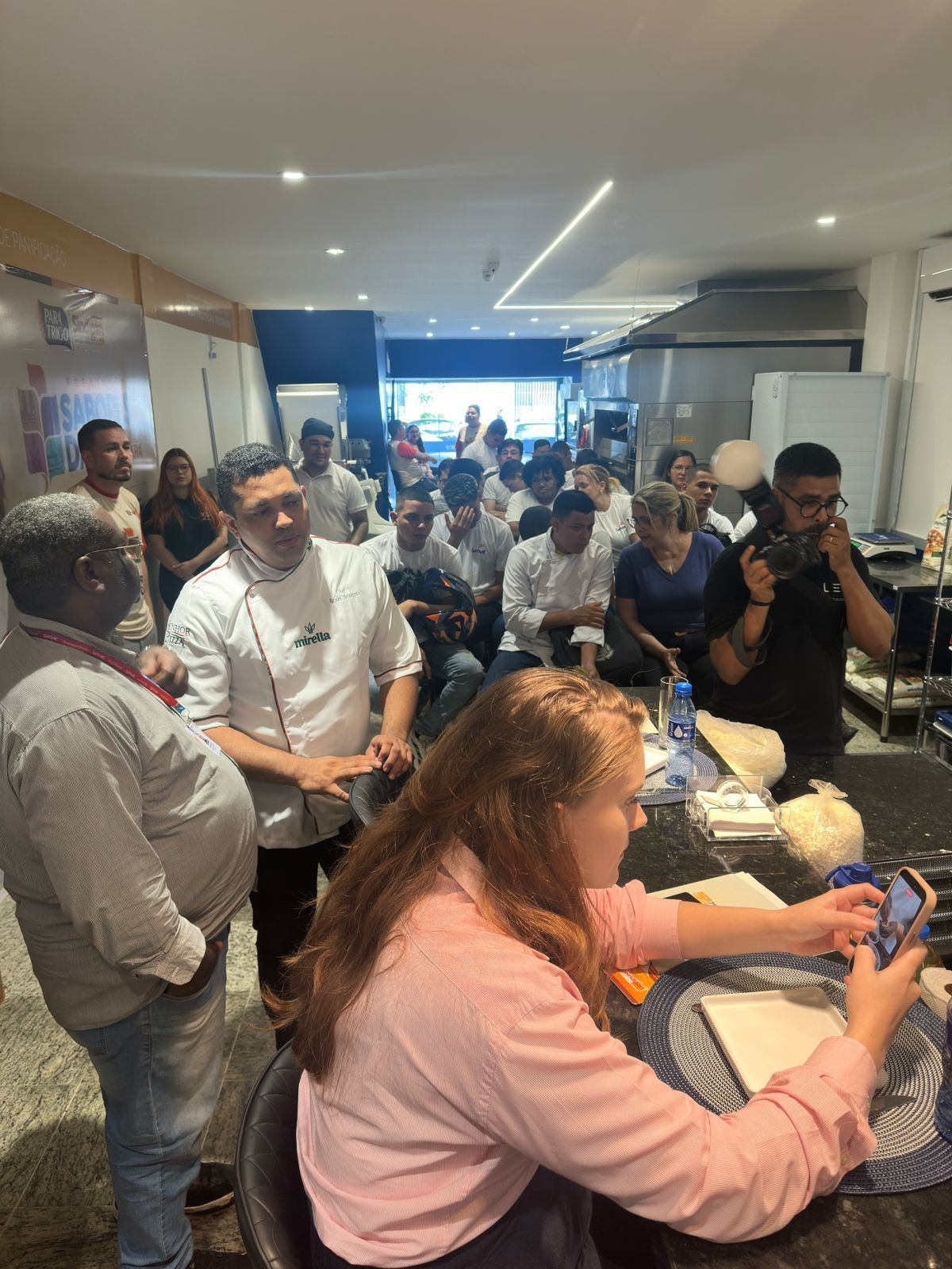Chef fazendo uma demonstração para uma multidão em uma cozinha, mulher tirando uma selfie.
