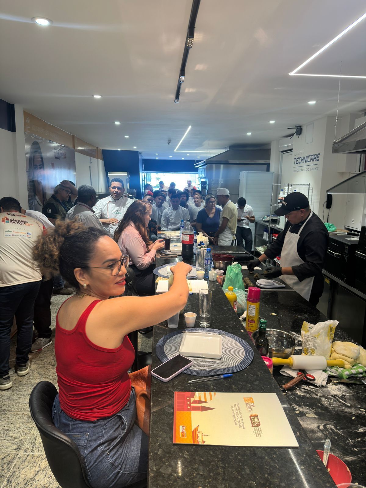 Uma cena de cozinha movimentada. Uma mulher de vermelho estende a mão em direção ao balcão. Pessoas estão comendo e cozinhando.