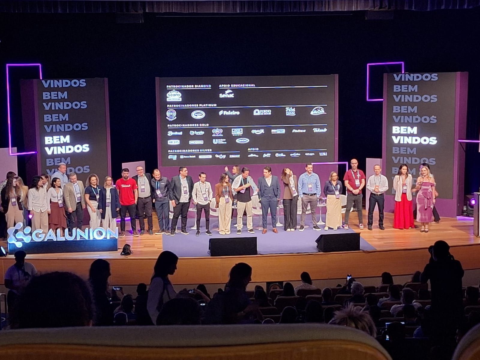 Grupo de pessoas no palco de um evento. Texto 