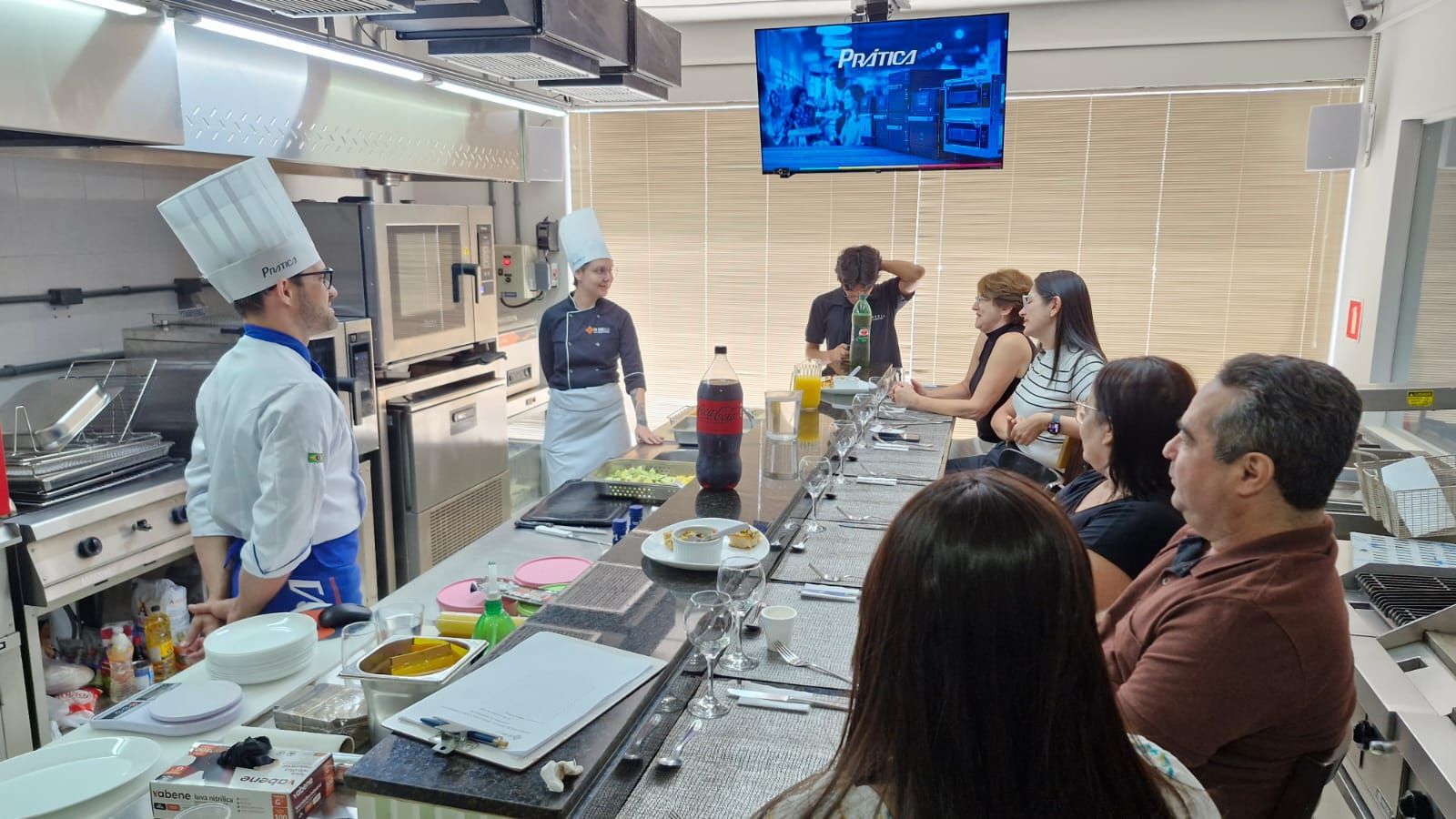 Chef dá aula de culinária; alunos observam no balcão de uma cozinha moderna.