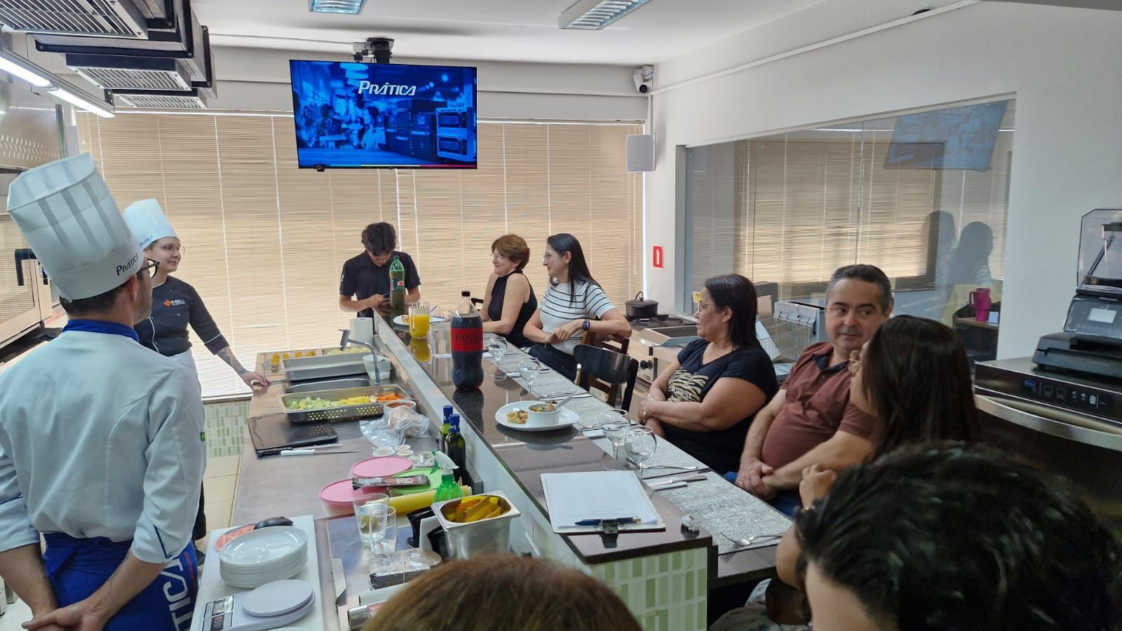 Chefs demonstram técnicas de culinária para um grupo de observadores sentados em uma cozinha moderna com TV.