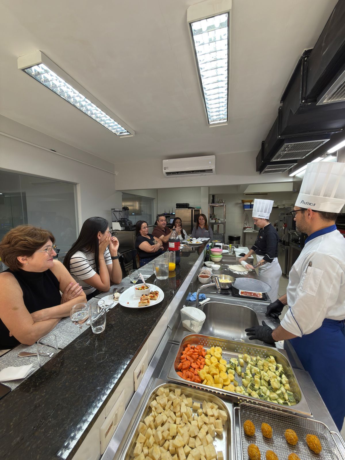 Chef demonstra técnicas culinárias para um grupo sentado em uma cozinha. Ingredientes e alimentos preparados estão em exposição.