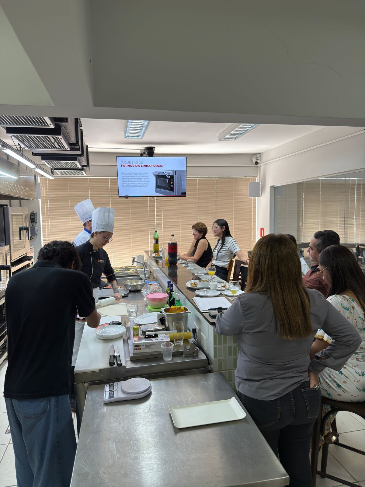 Chef demonstrando culinária para um grupo em uma cozinha com tela de TV.