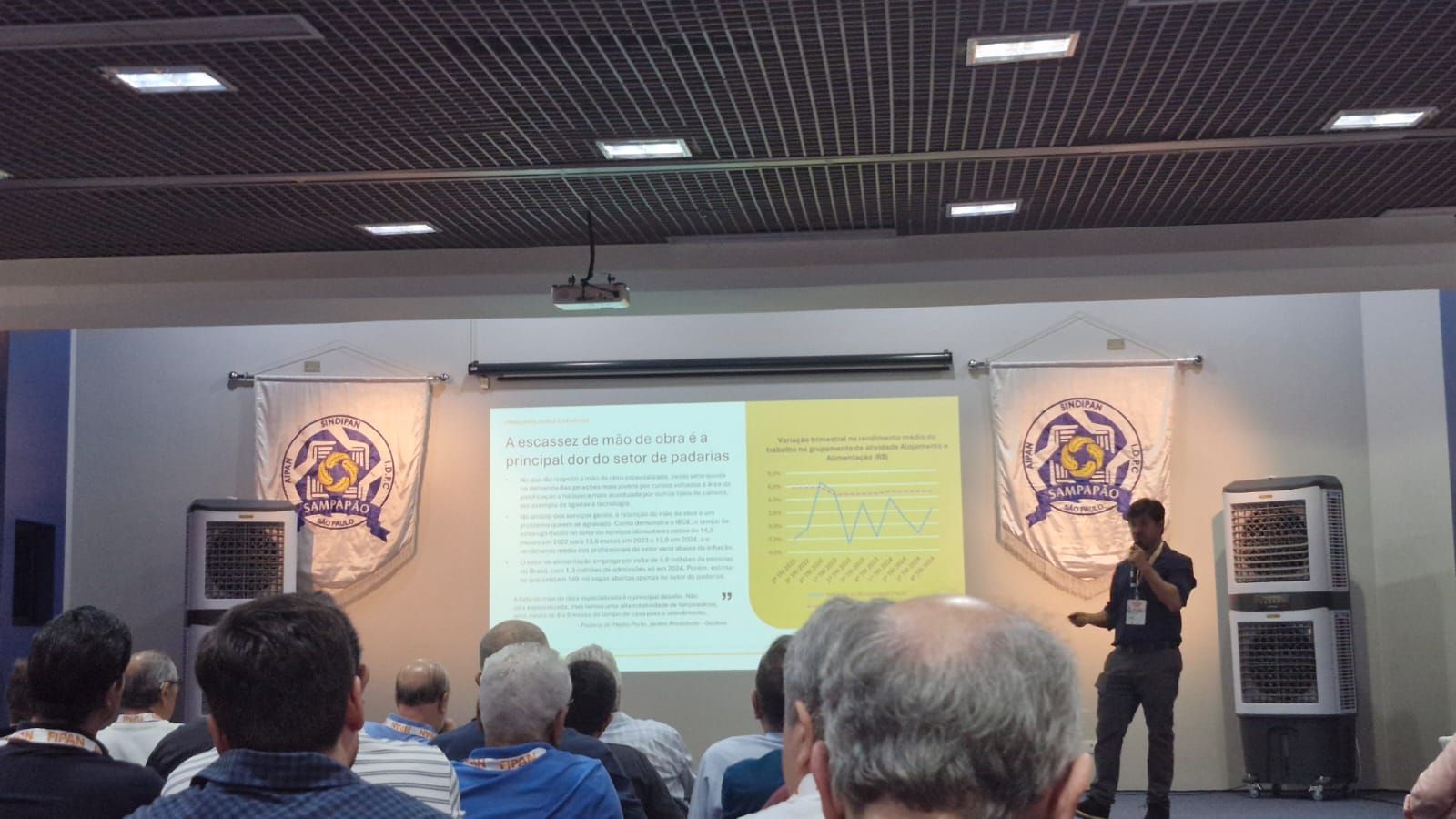 Um homem fazendo uma apresentação em uma sala de conferências com público e slides.